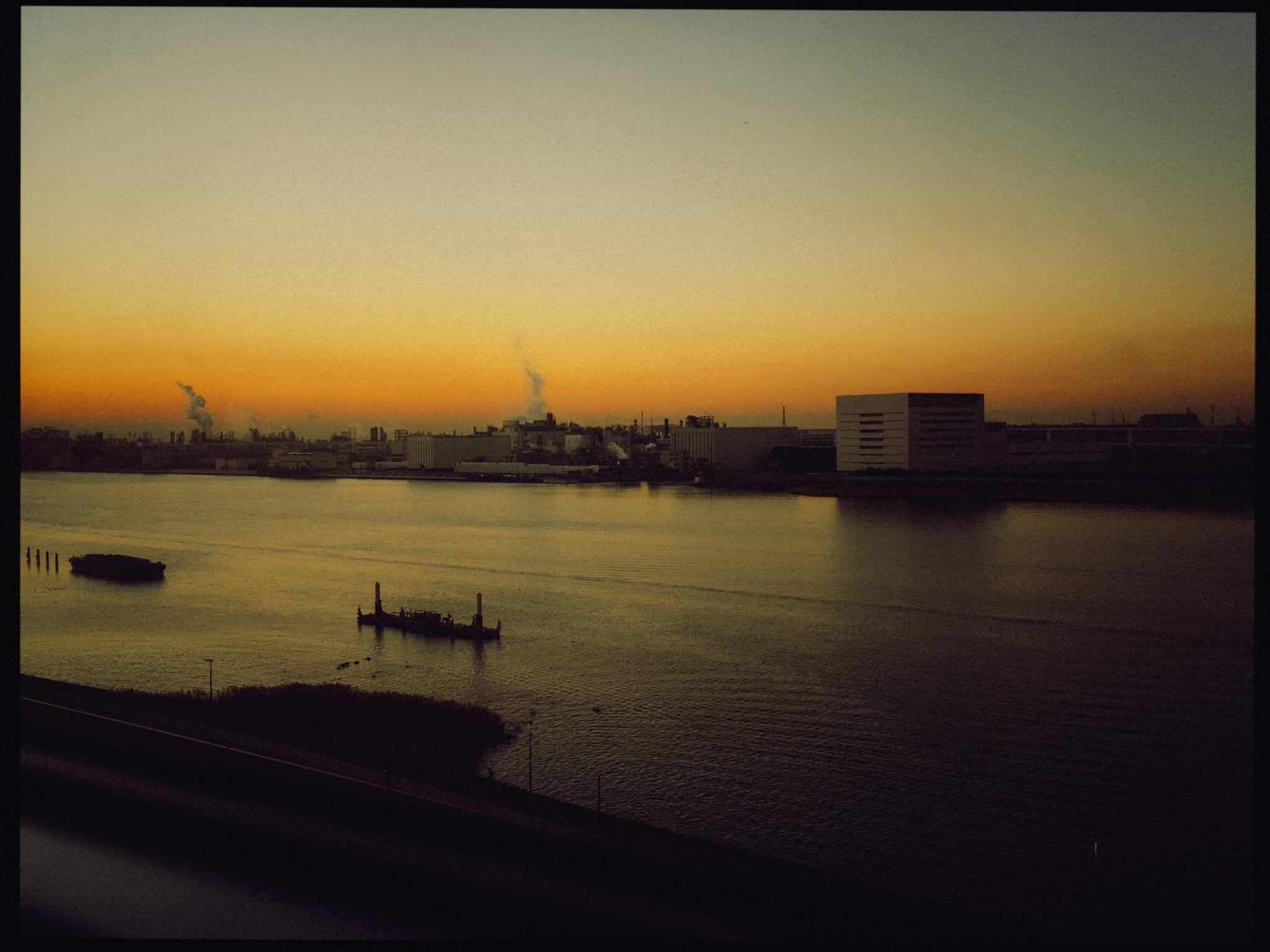 Small boats rest on still water beneath a yellow sunset and a distant industrial skyline.