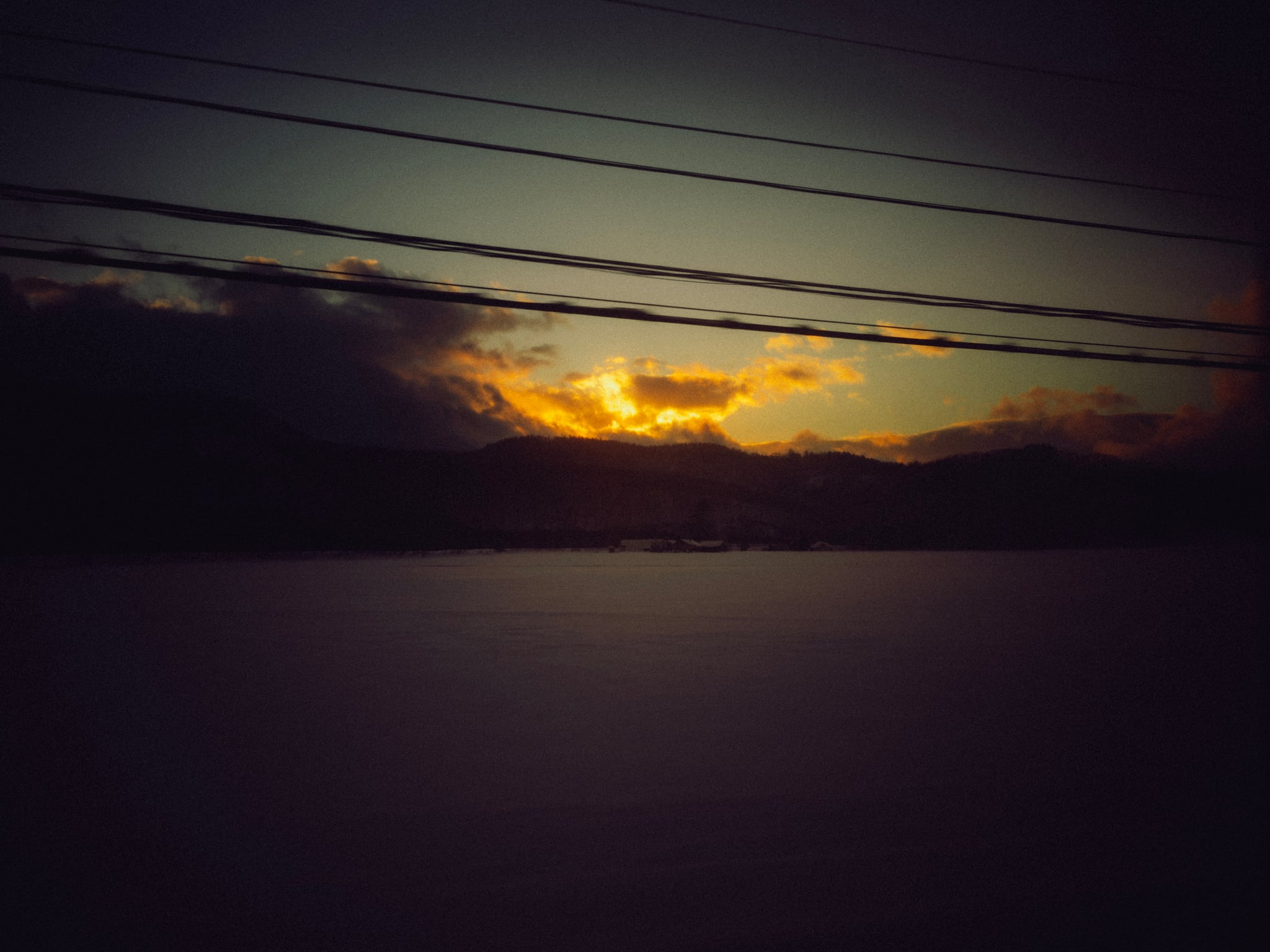 An orange opening in dark sunset clouds above water, crossed by overhead wires.