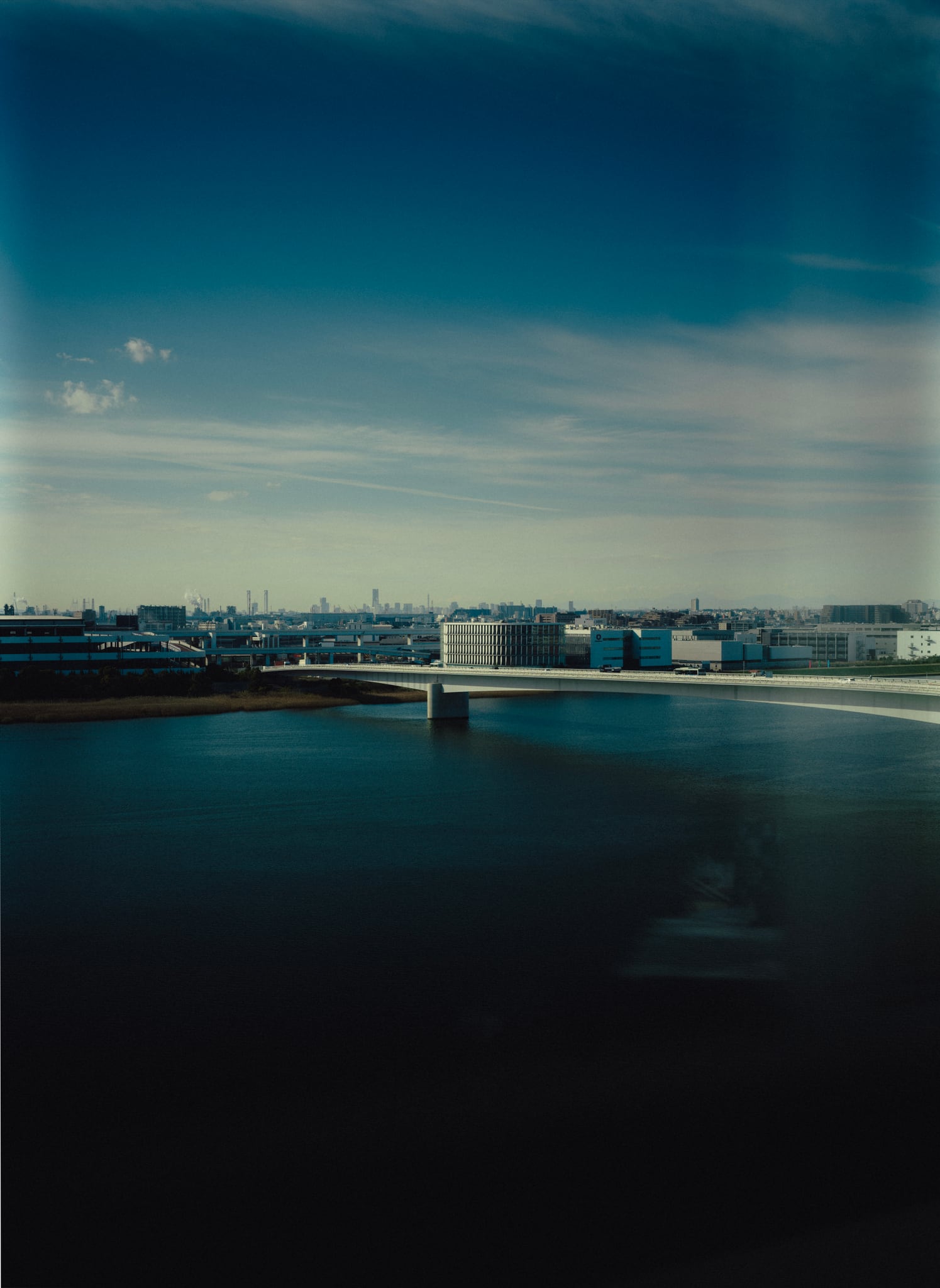 A pale bridge stretches across blue water toward a distant city under a clear sky.