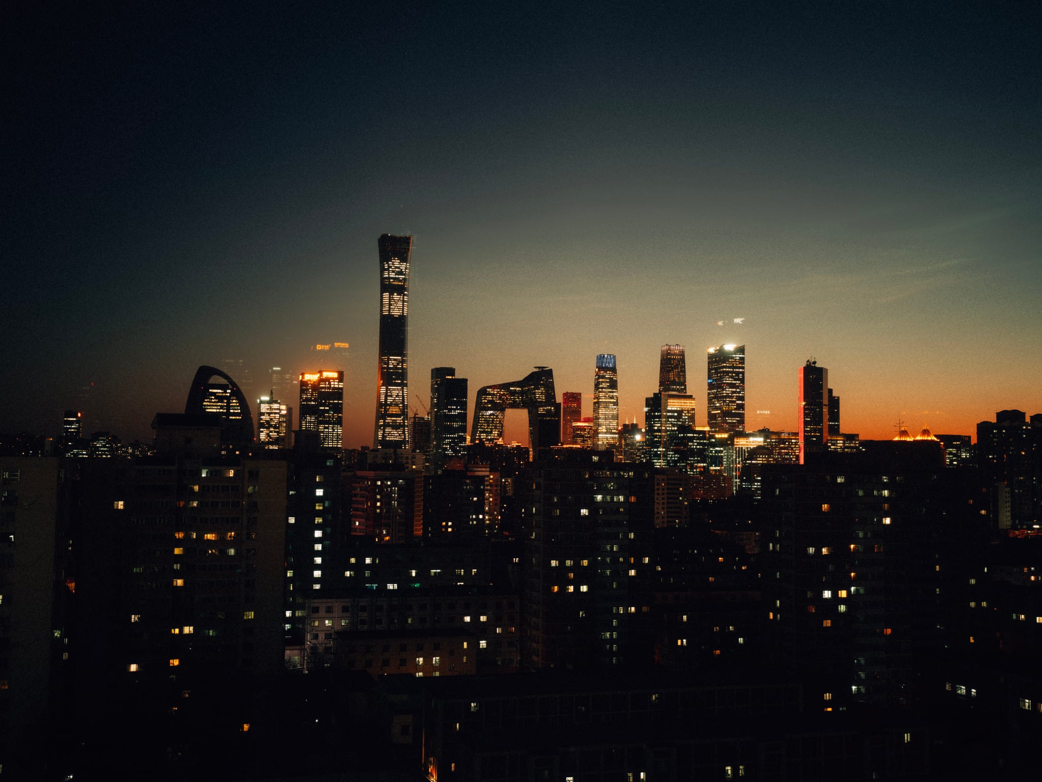 Illuminated towers rise above dark apartment buildings against the last orange light of sunset.