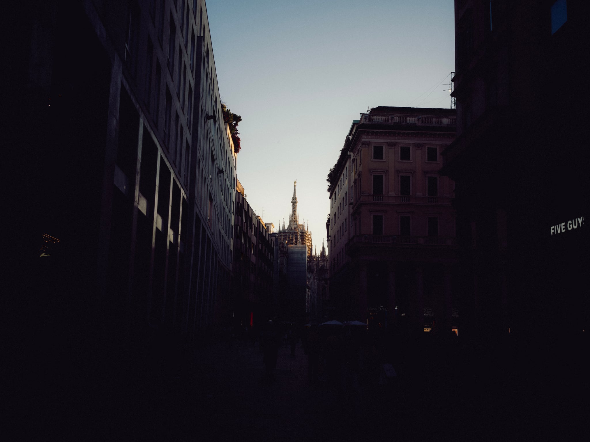 A distant spire stands between tall buildings along a narrow, shadowed city street.