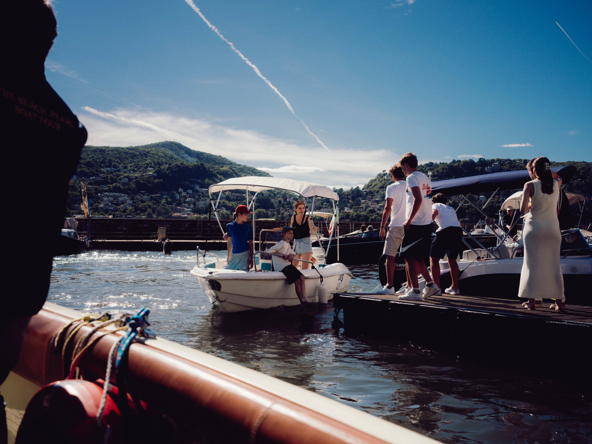 People stand beside a small boat at a lakeside jetty, with mountains beyond the water.