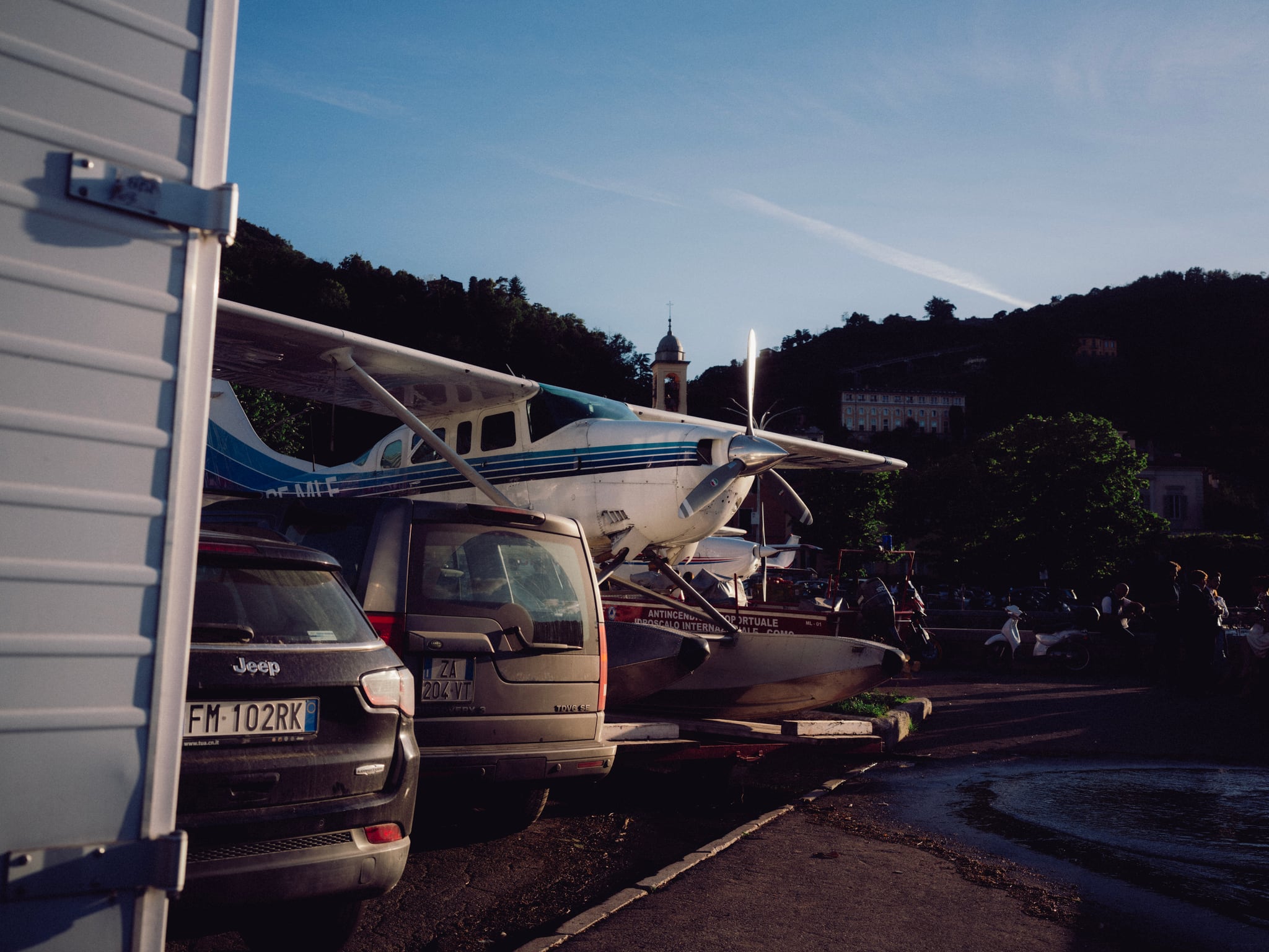 Parked cars line a narrow space beside buildings near a wooded hillside.