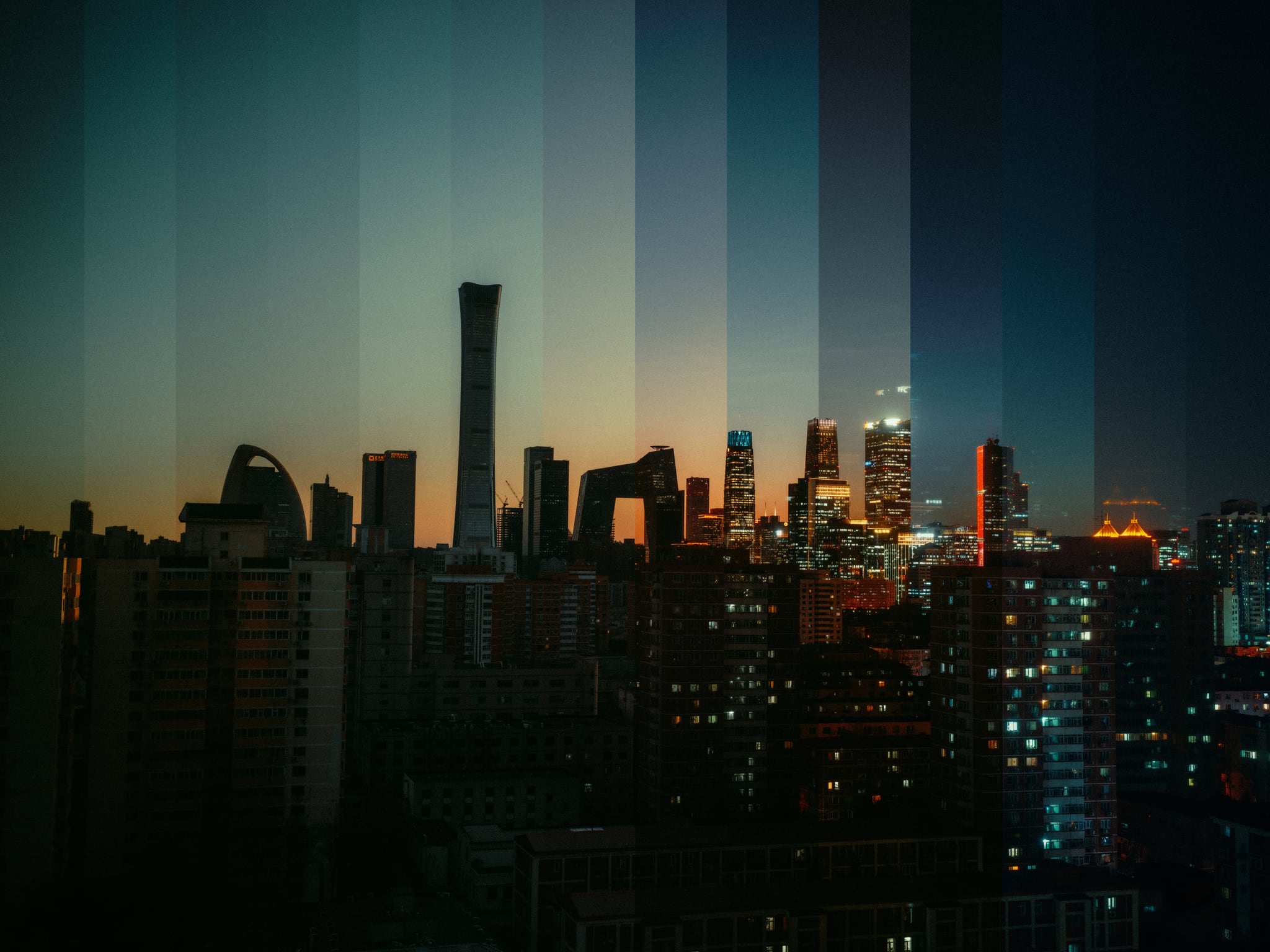 Vertical bands of changing light divide a city skyline at dusk above dark apartment buildings.