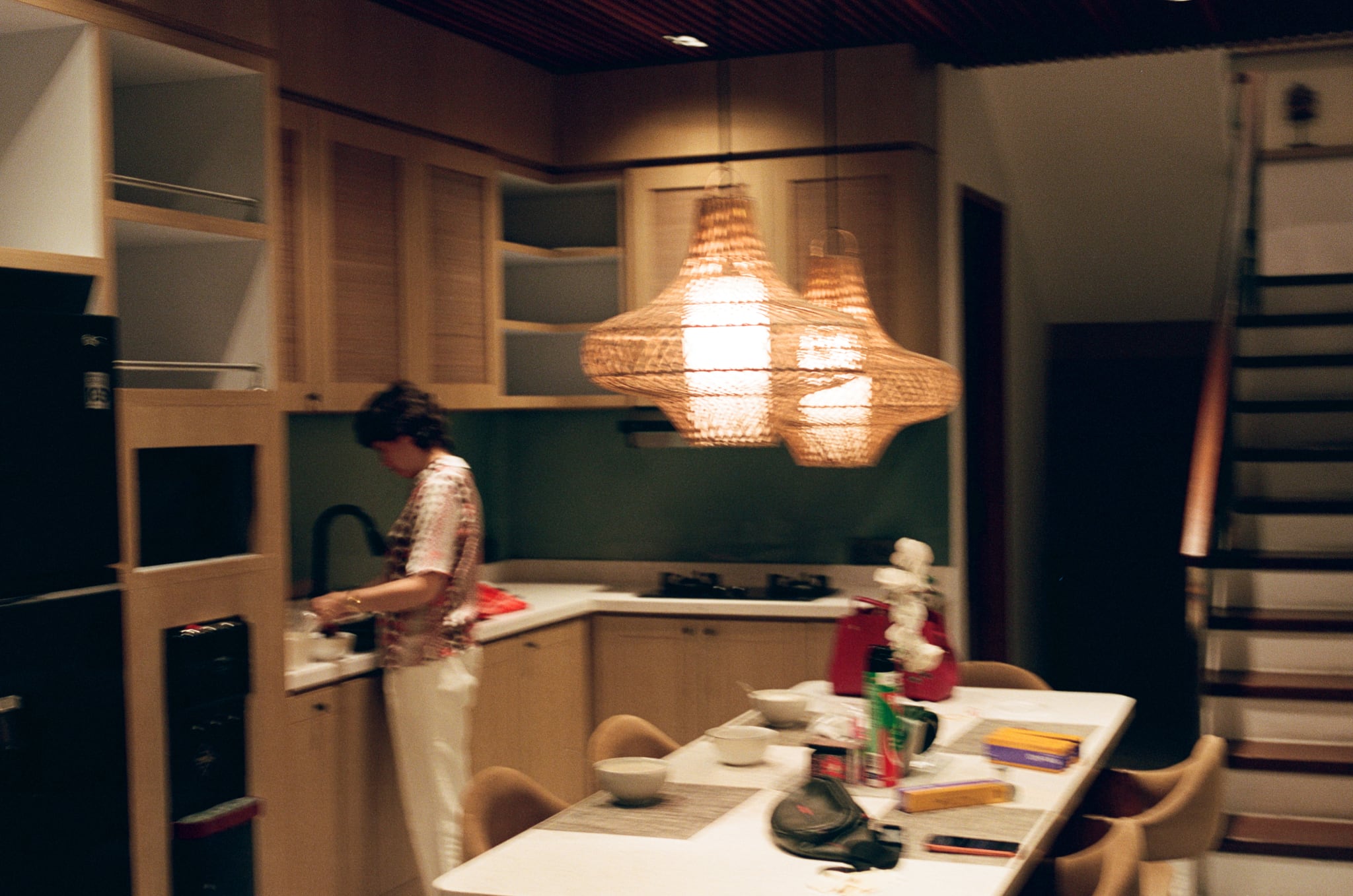 A person stands in a kitchen beneath a glowing woven pendant light beside a dining table.