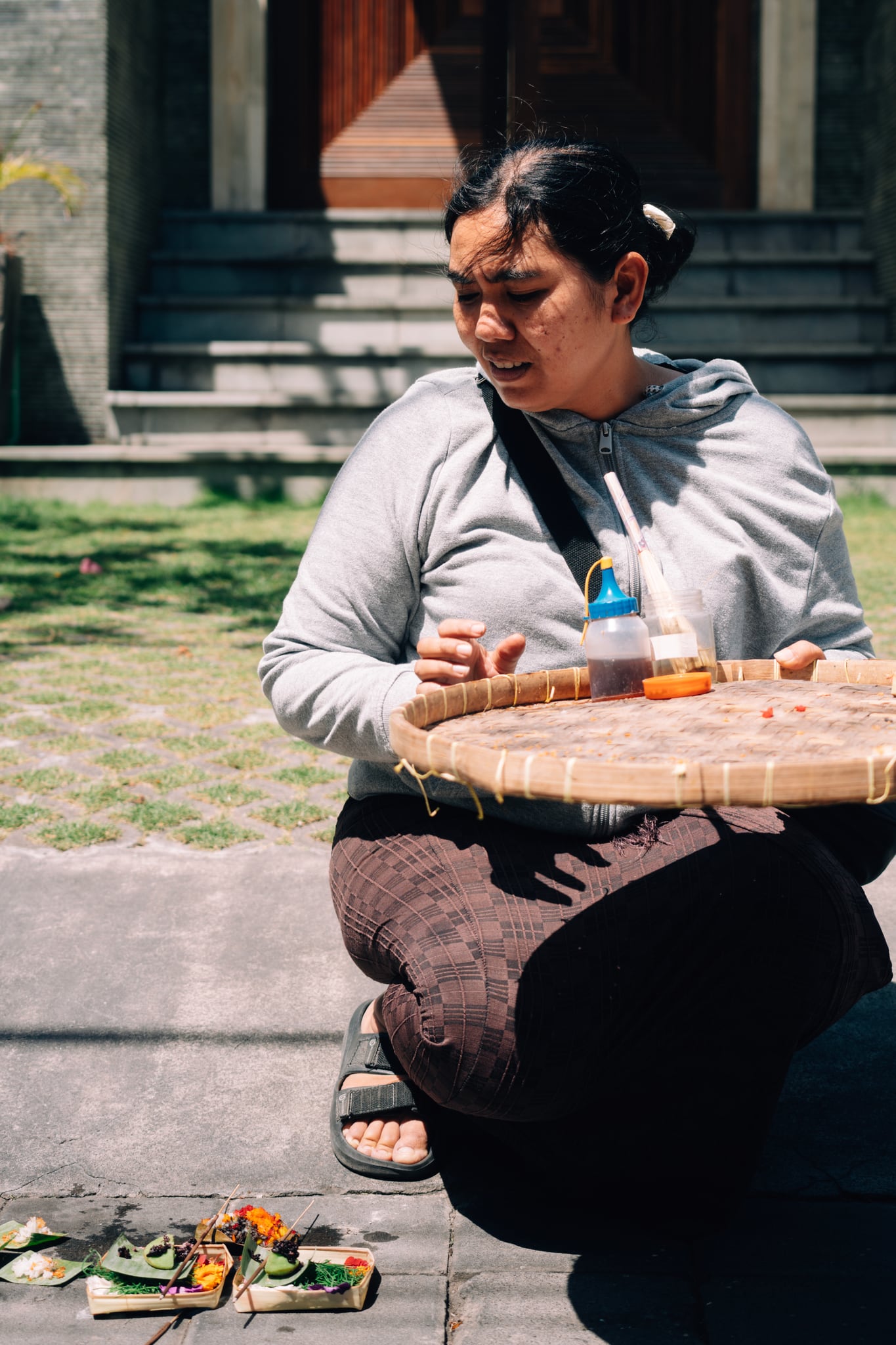 A person sits in sunlight with a round woven tray on their lap in a Bali courtyard.