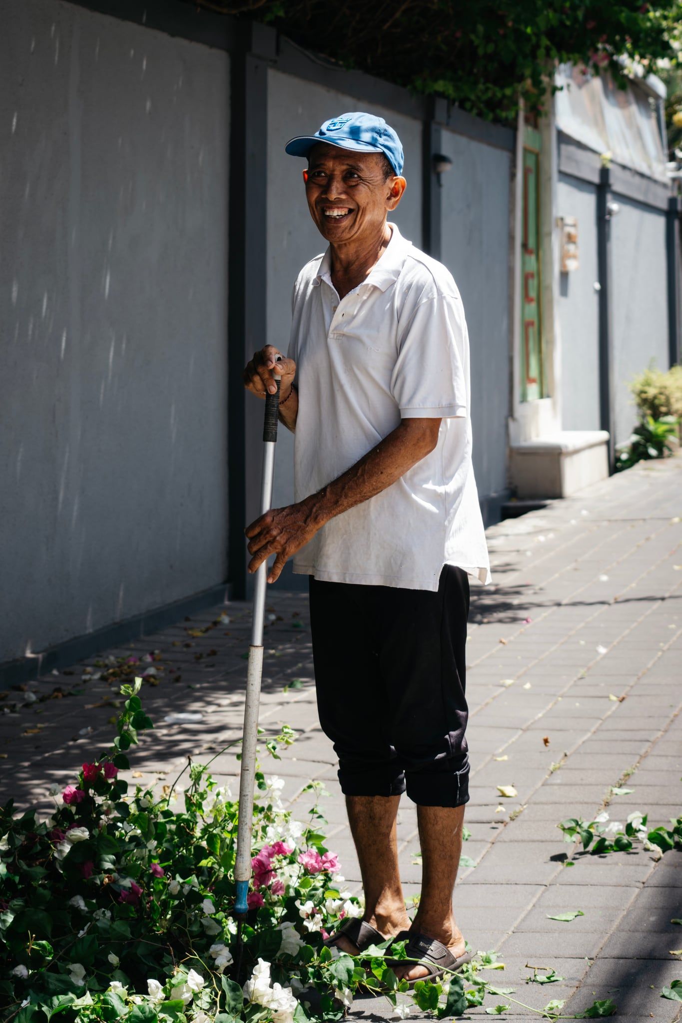 A person in a pale shirt and blue cap stands beside a wall and low plants in Bali.