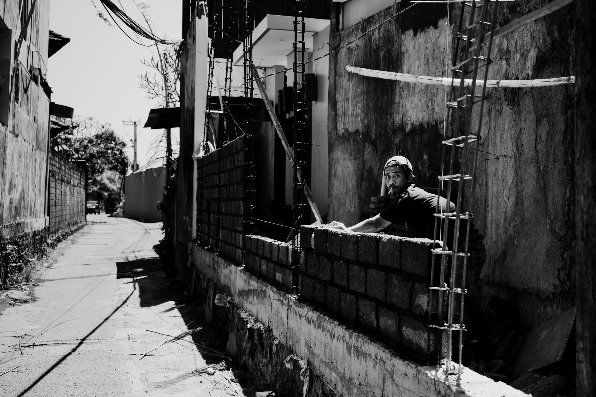 A black-and-white photograph of a narrow Bali lane, with a person beside a low wall in strong sunlight.