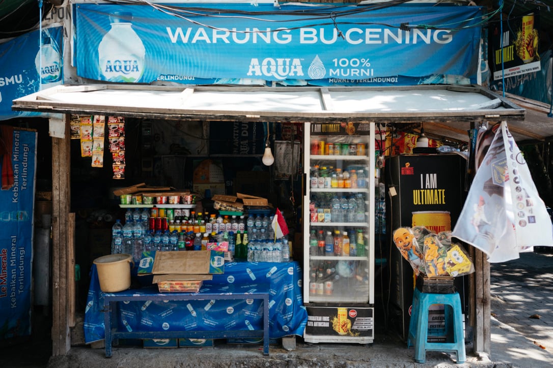 A small Bali shop beneath a blue Warung Bu Cening sign displays drinks, goods, and a refrigerator.