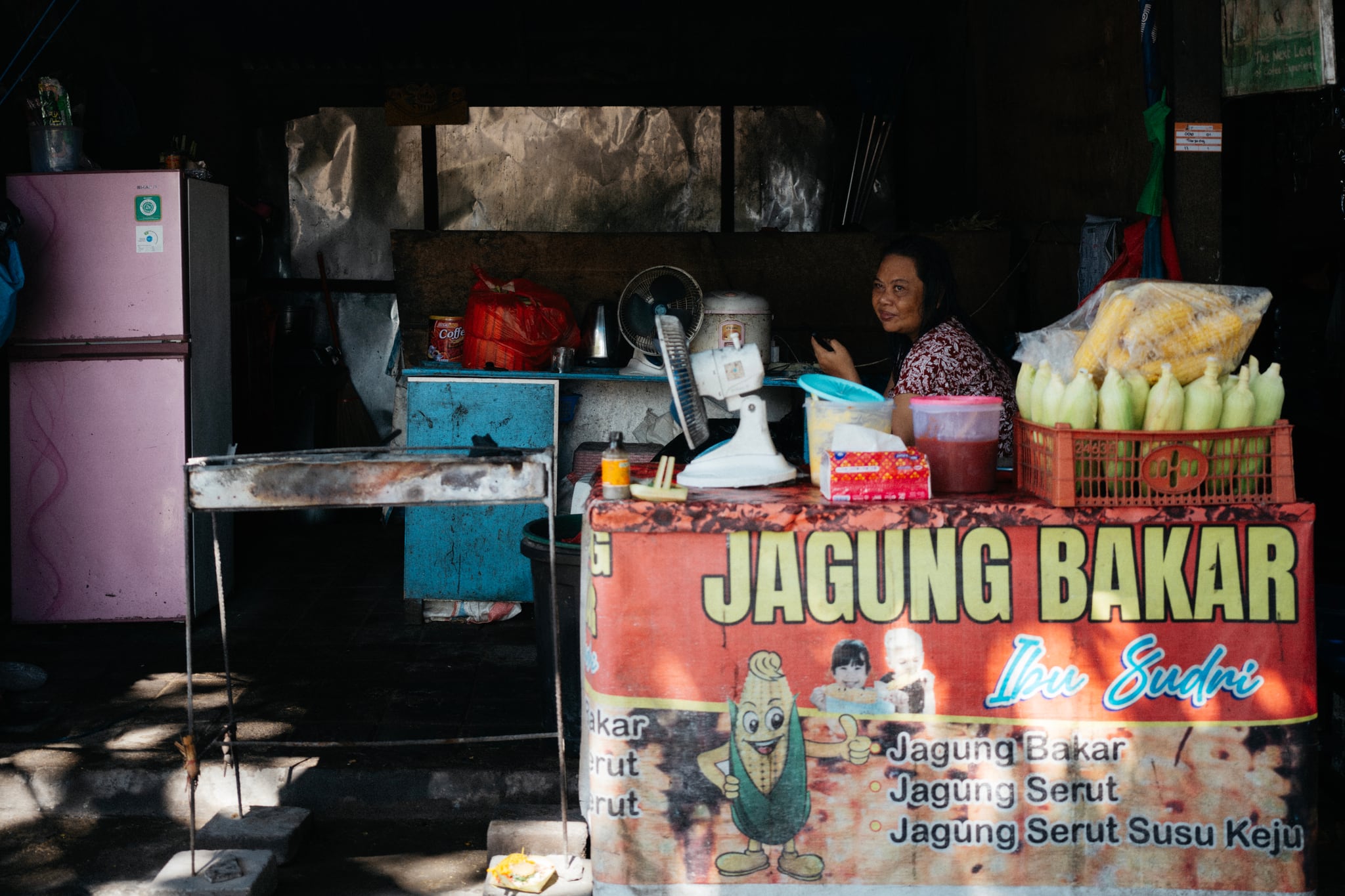 A person sits behind a counter of corn and containers inside a dimly lit Bali food stall.
