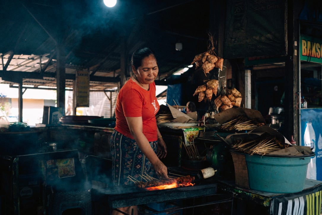 A person in a red shirt tends food over glowing coals inside a shaded Bali stall.