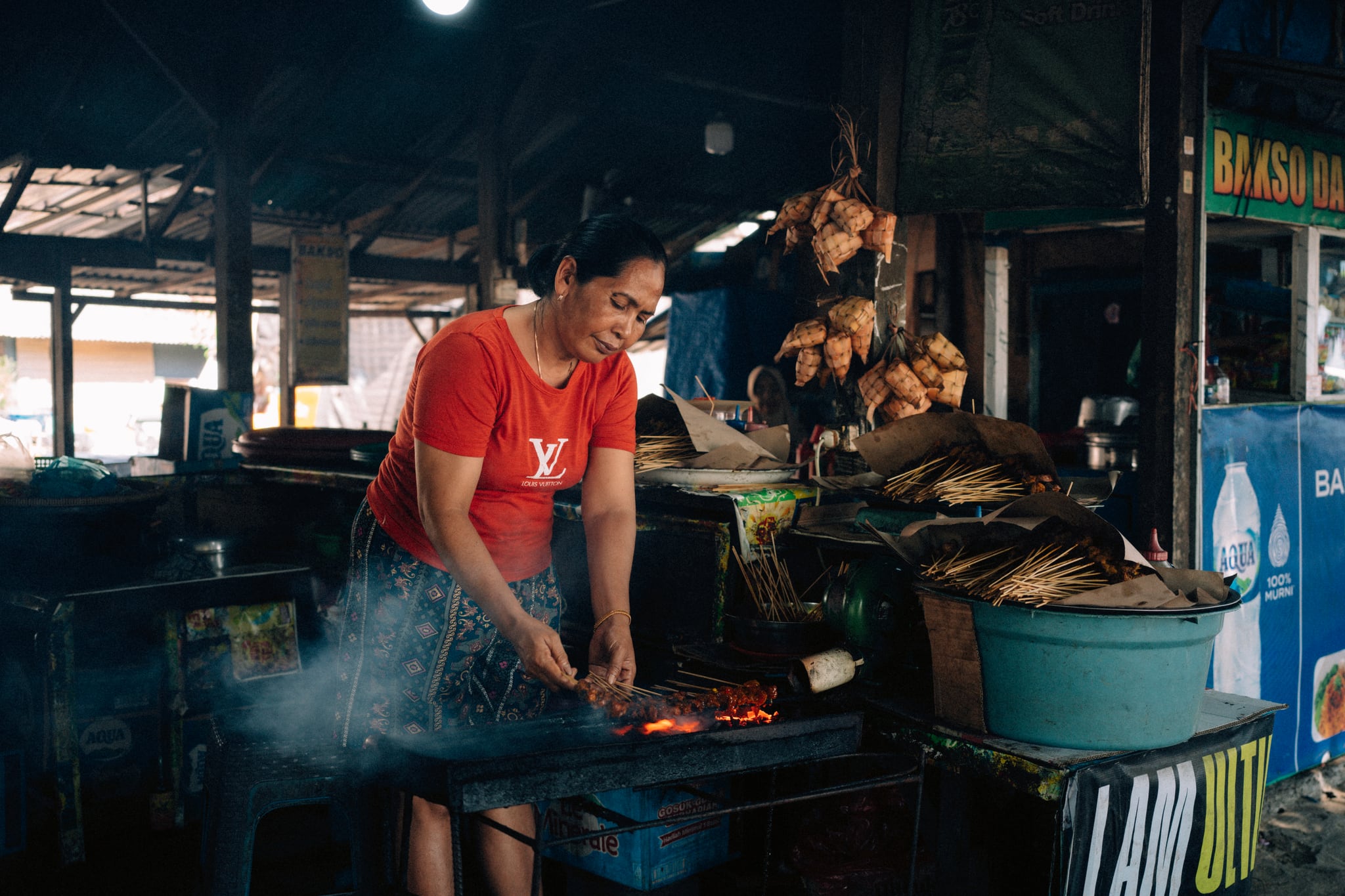 A cook in a red shirt leans over a glowing grill beneath the dark roof of a Bali food stall.
