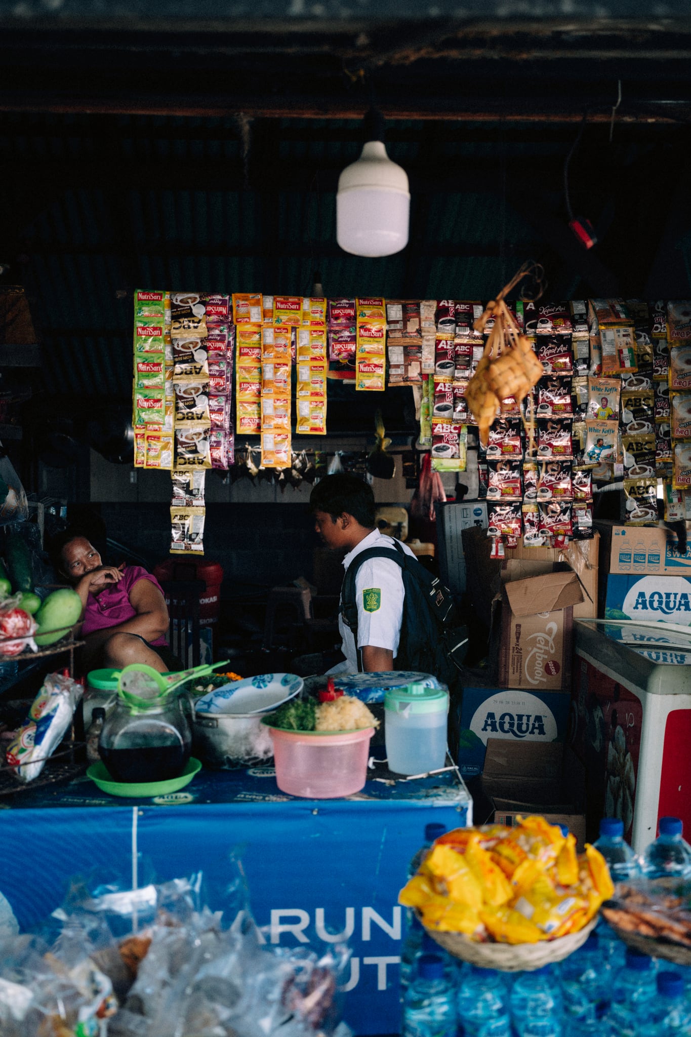 Rows of hanging packets and containers fill the shaded interior of a roadside shop in Bali.
