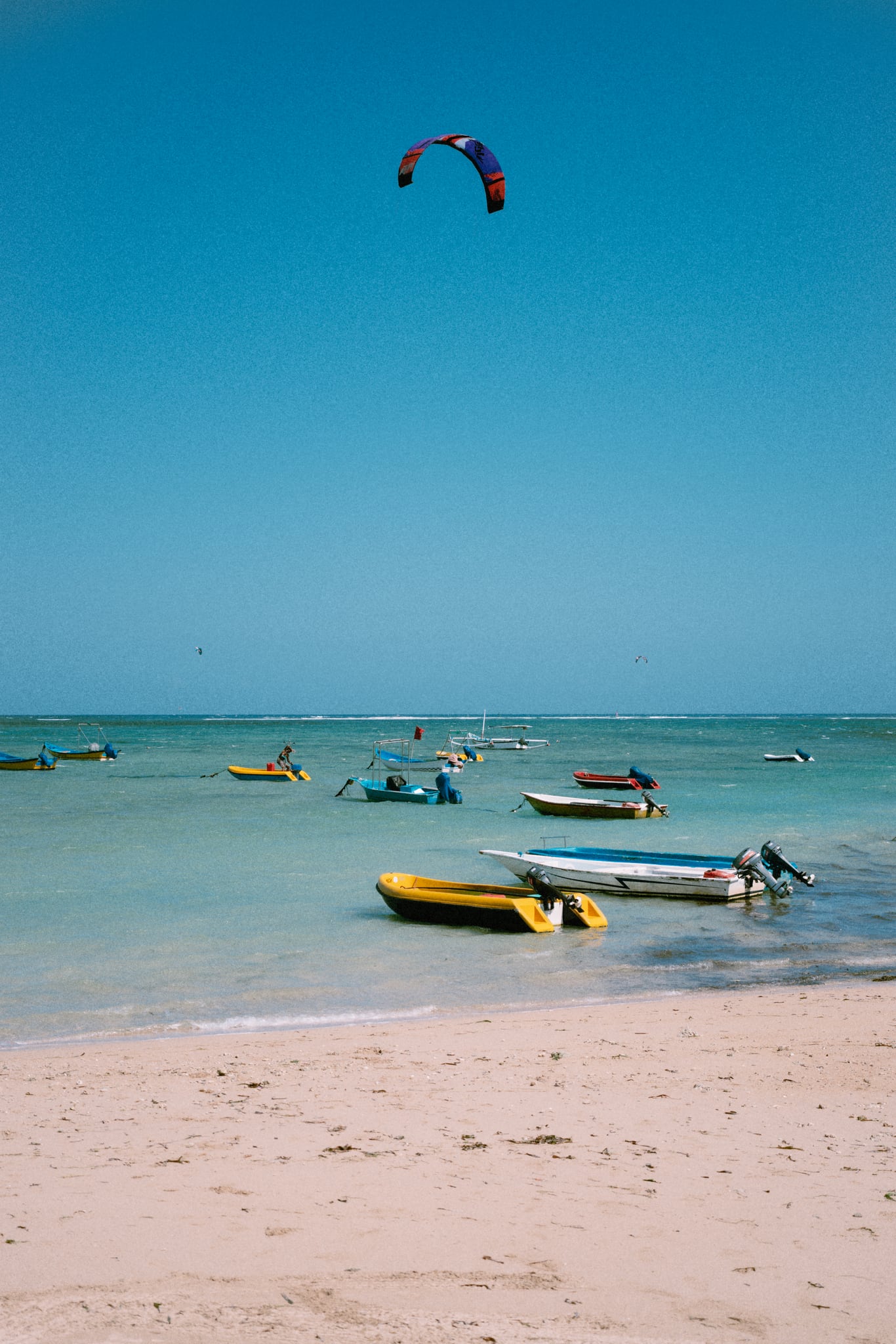 Small boats float beside a pale sandy Bali shoreline, with a kite high overhead.