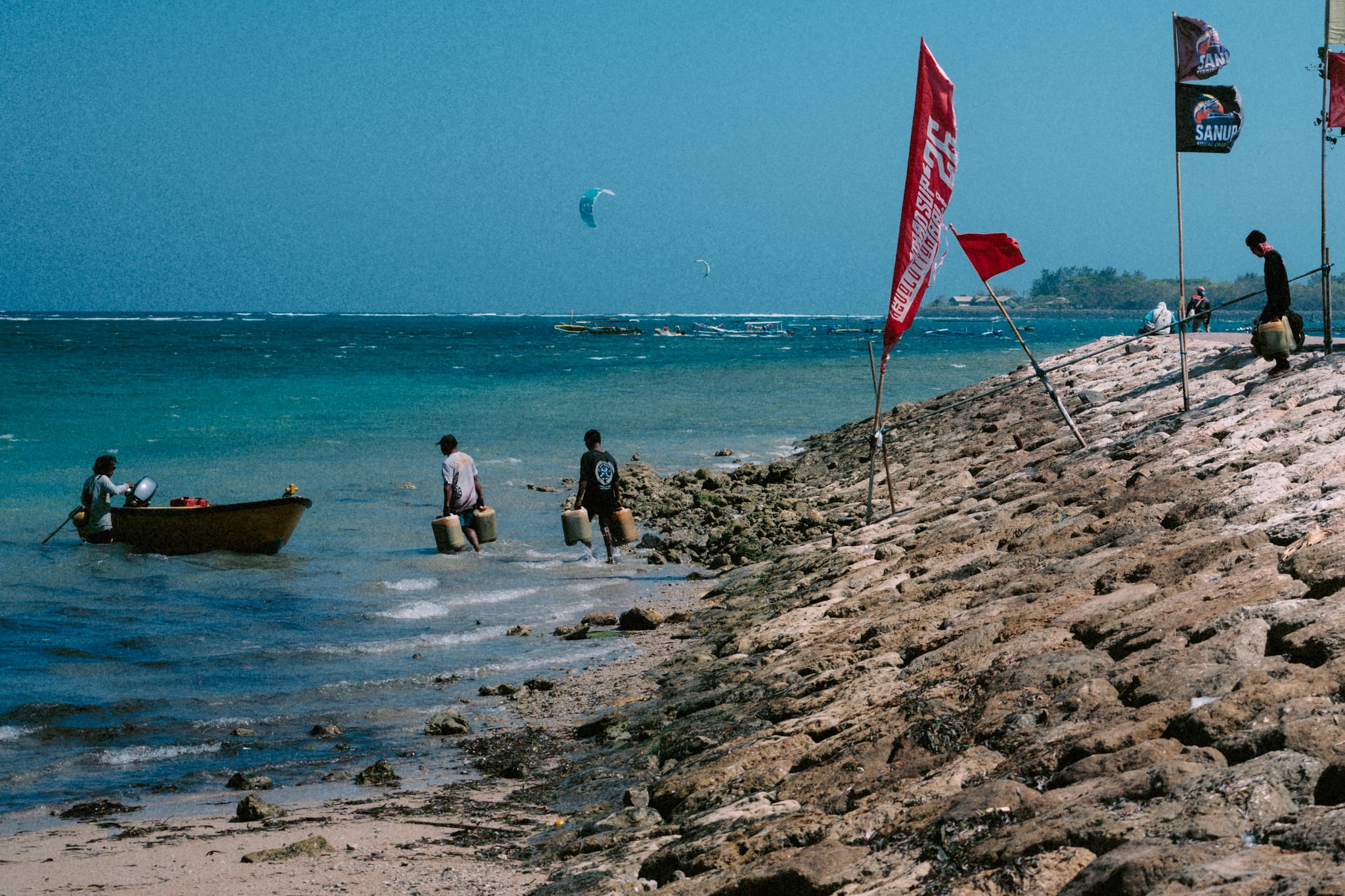 People and a small boat gather at the water below flags along a stone-lined Bali beach.