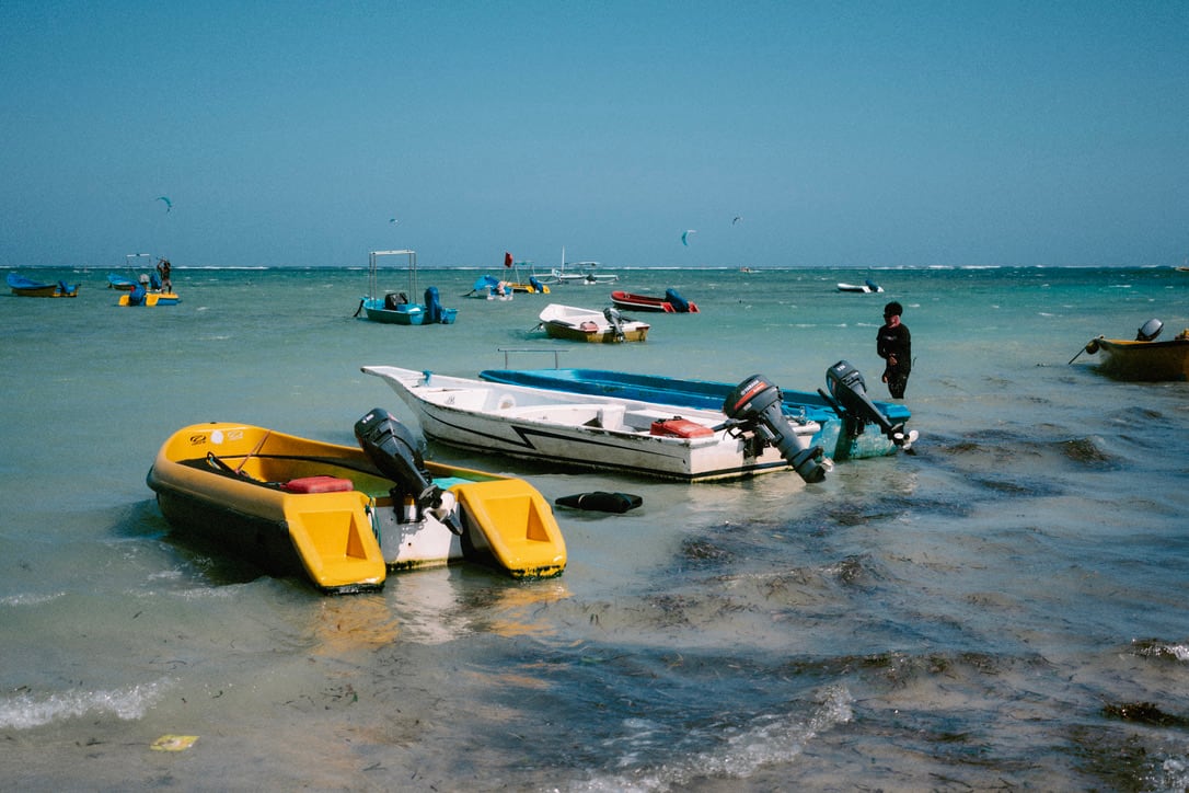 Yellow and white boats point toward the shore as a person stands in the shallow sea in Bali.