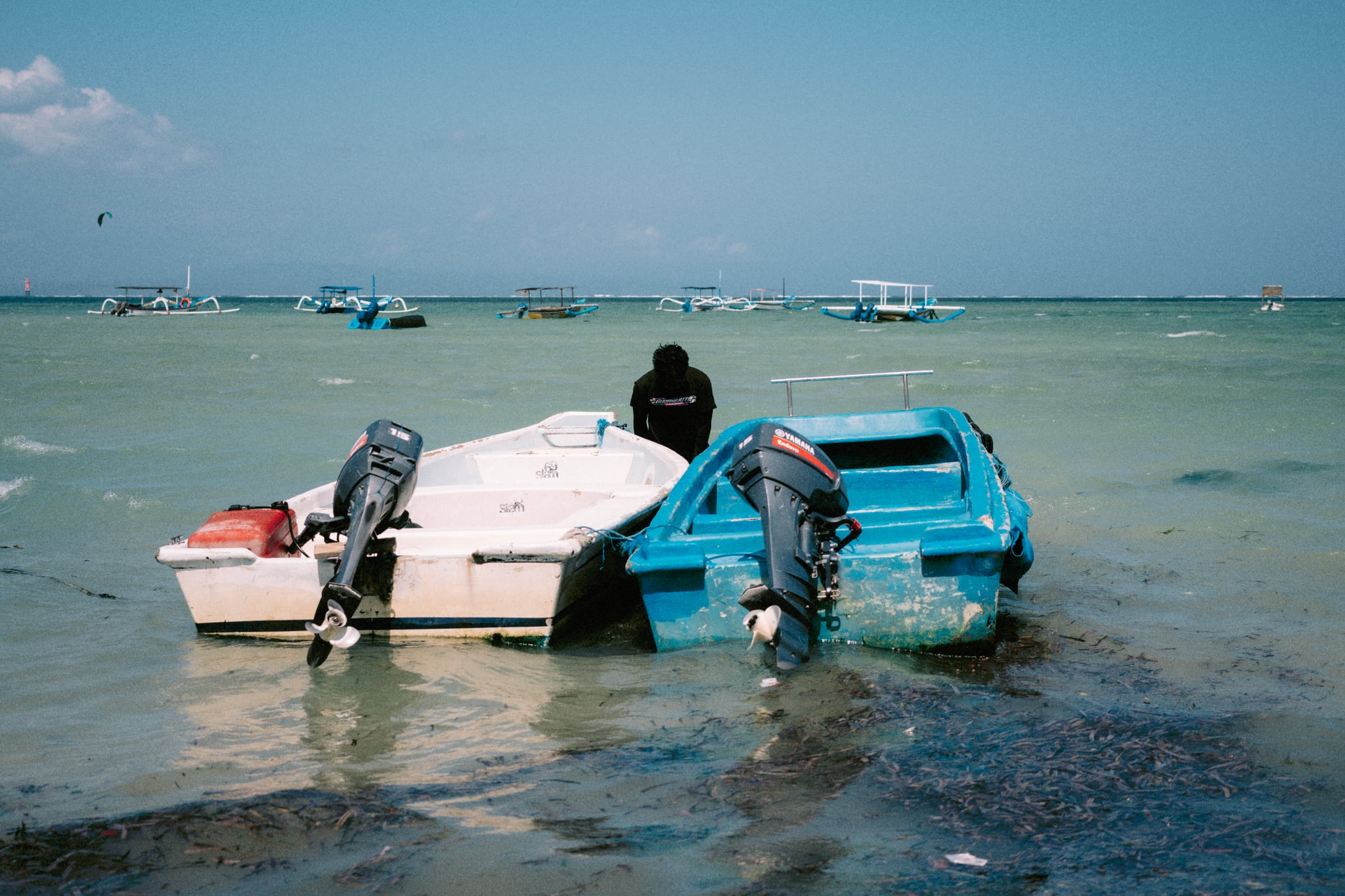 A white boat and a blue boat sit side by side in pale shallow water beside a person in Bali.