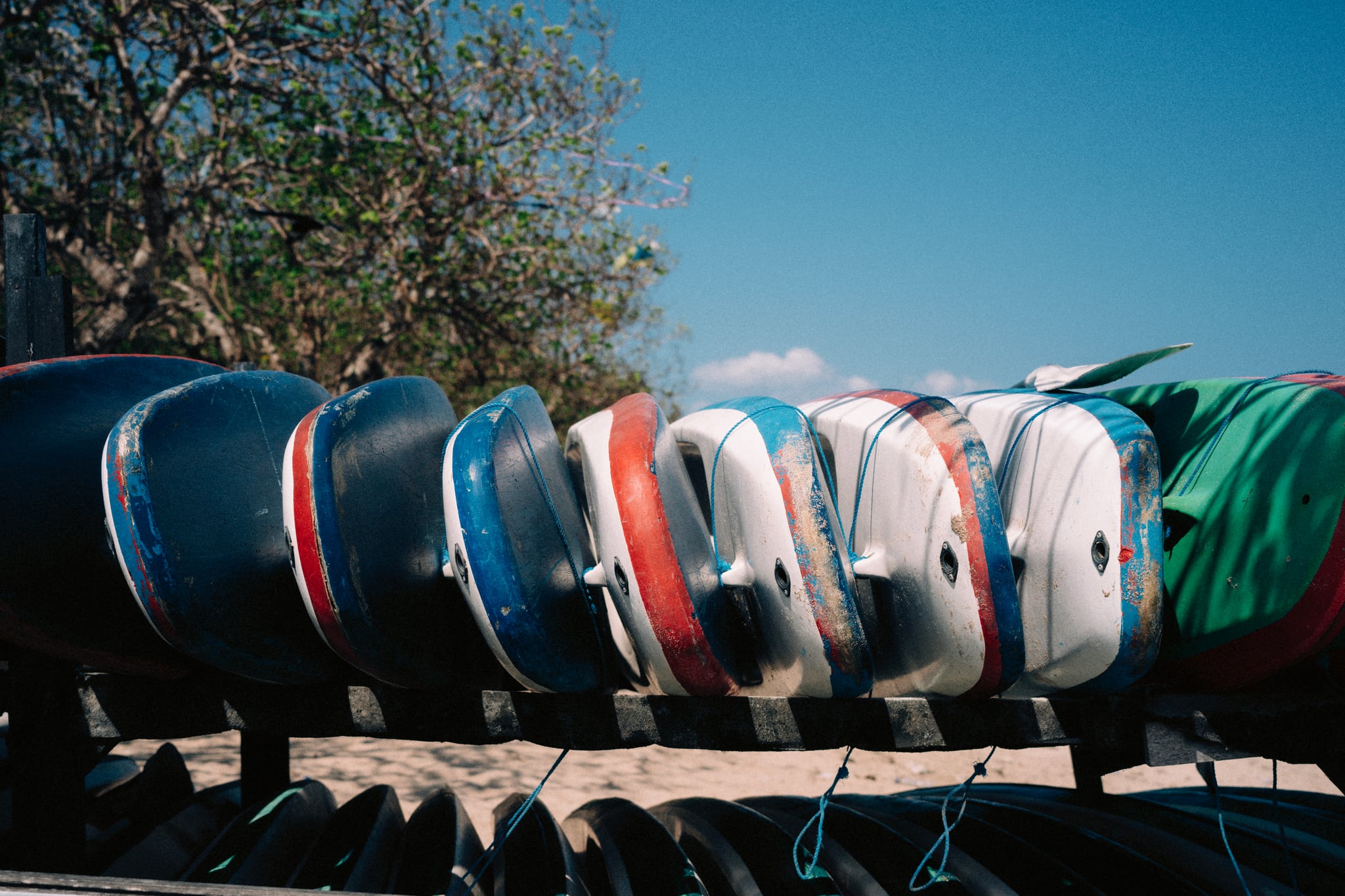 A row of colourful boards is stacked upright beneath leafy trees near a Bali beach.