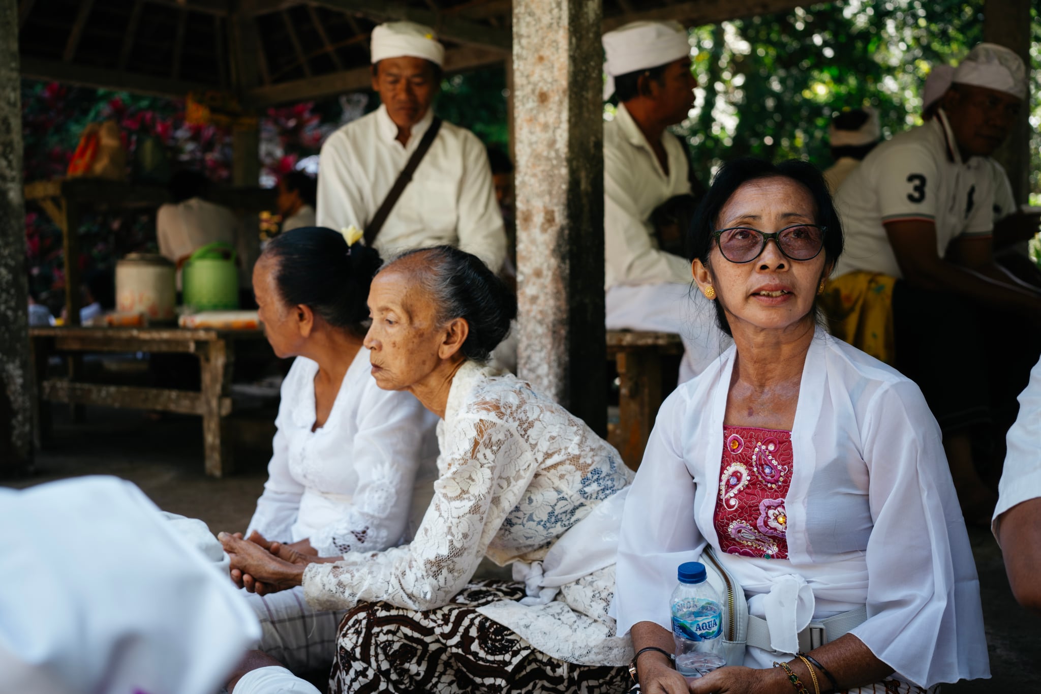 A group in pale clothing sits together in the shade of a wooden pavilion in Bali.