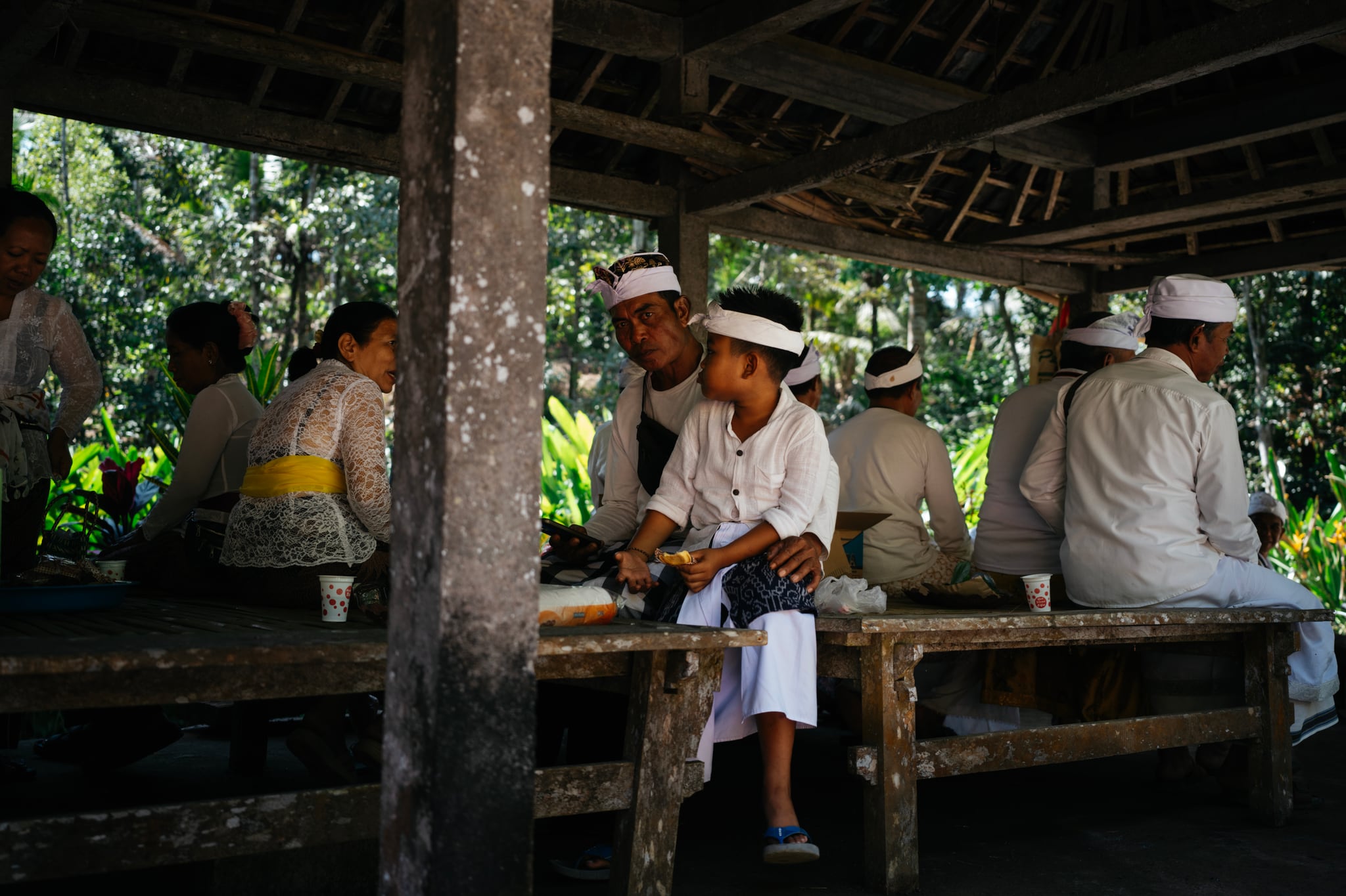 People gather around a table beneath a timber roof, with green foliage beyond, in Bali.