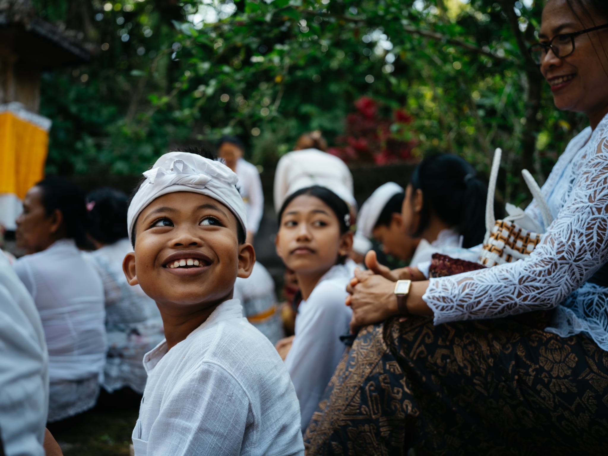 A smiling child in a white headwrap looks upward among a group of people in Bali.
