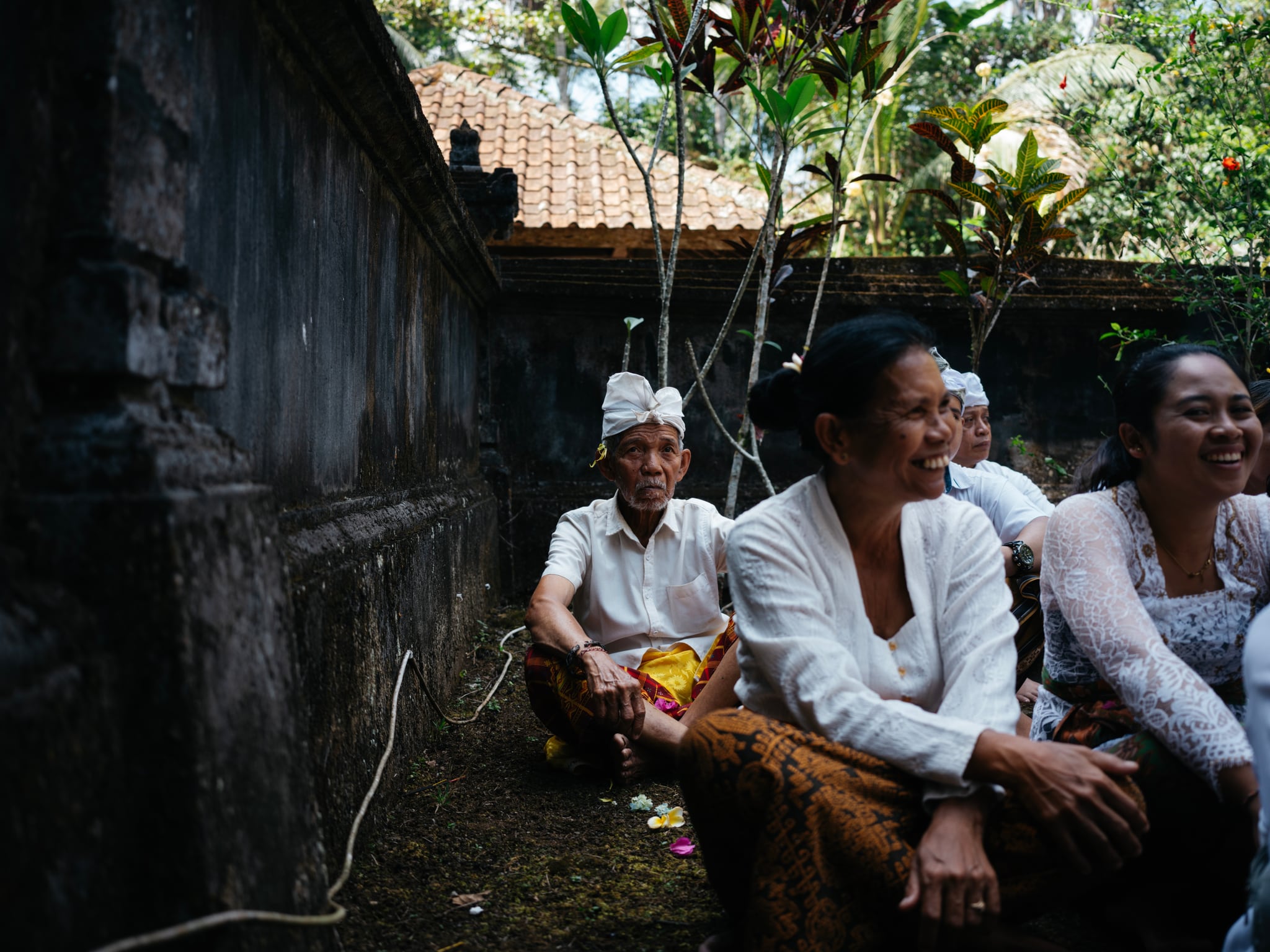 Seated people talk beside a rough wall and tropical plants in a Bali courtyard.
