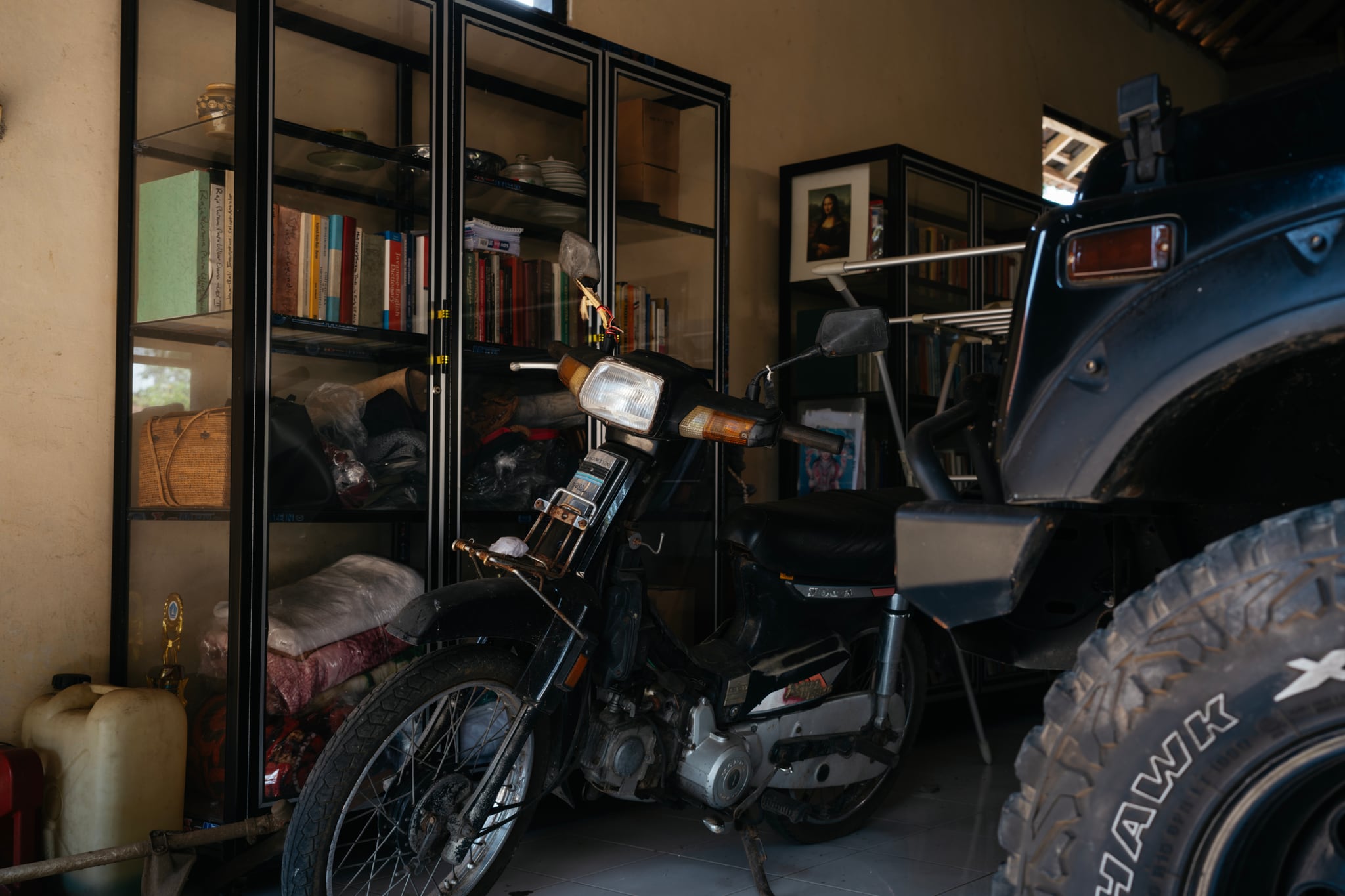 A motorcycle, a large vehicle tyre, and shelves of belongings fill a dim garage in Bali.