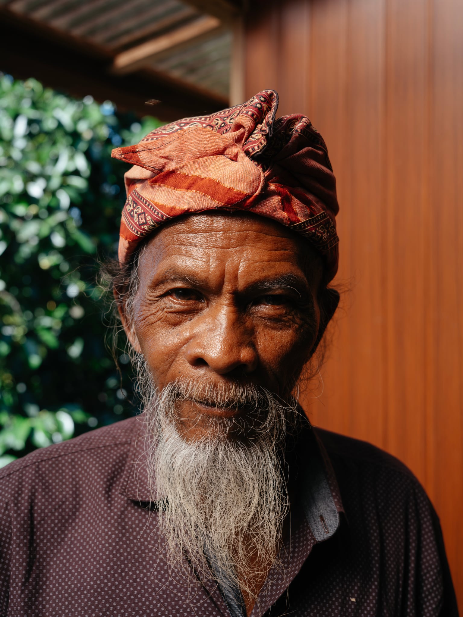 A person with a long grey beard and a red patterned headwrap faces the camera in Bali.