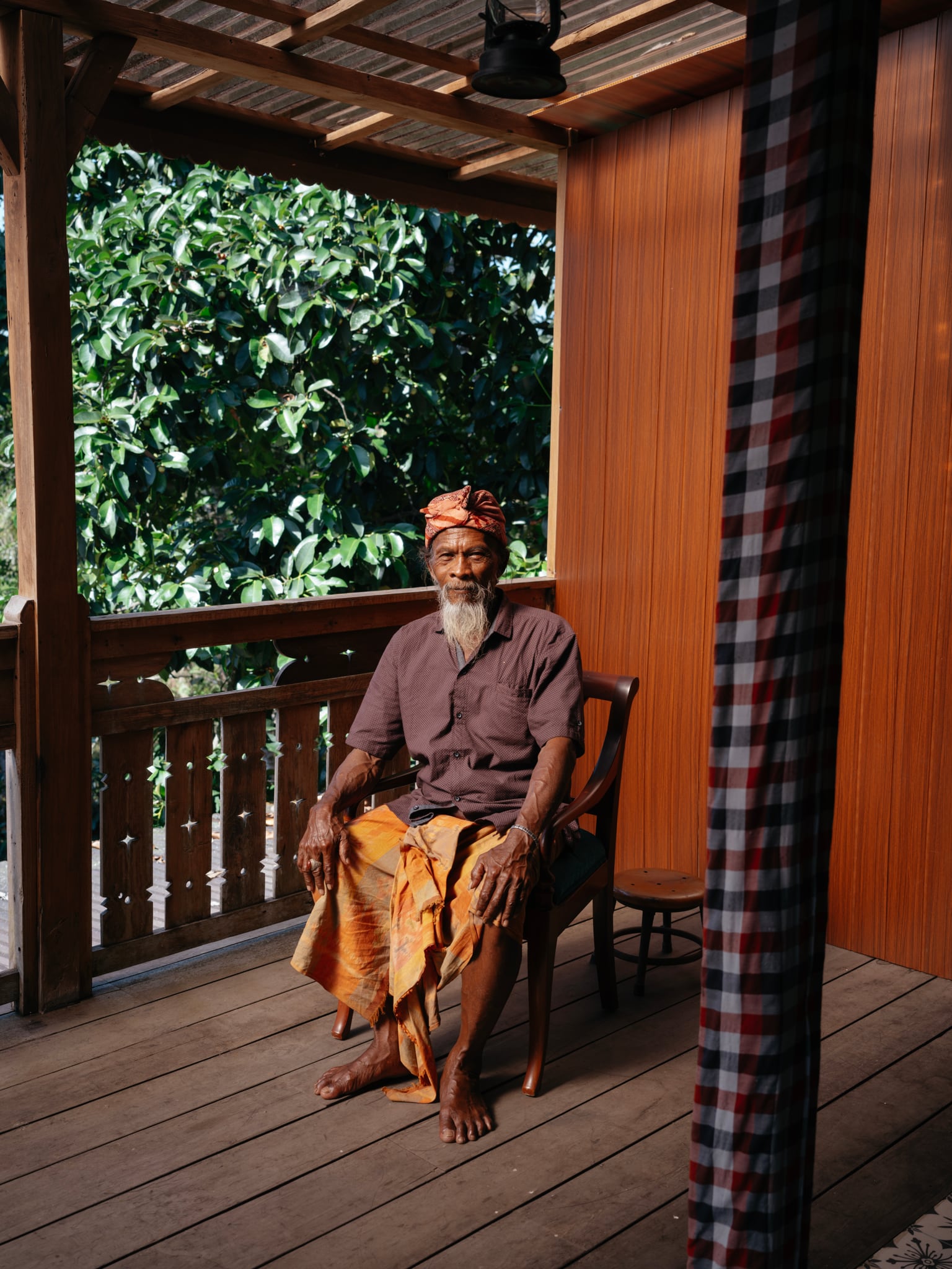 A person wearing a patterned wrap sits on a chair beside a doorway on a wooden veranda in Bali.