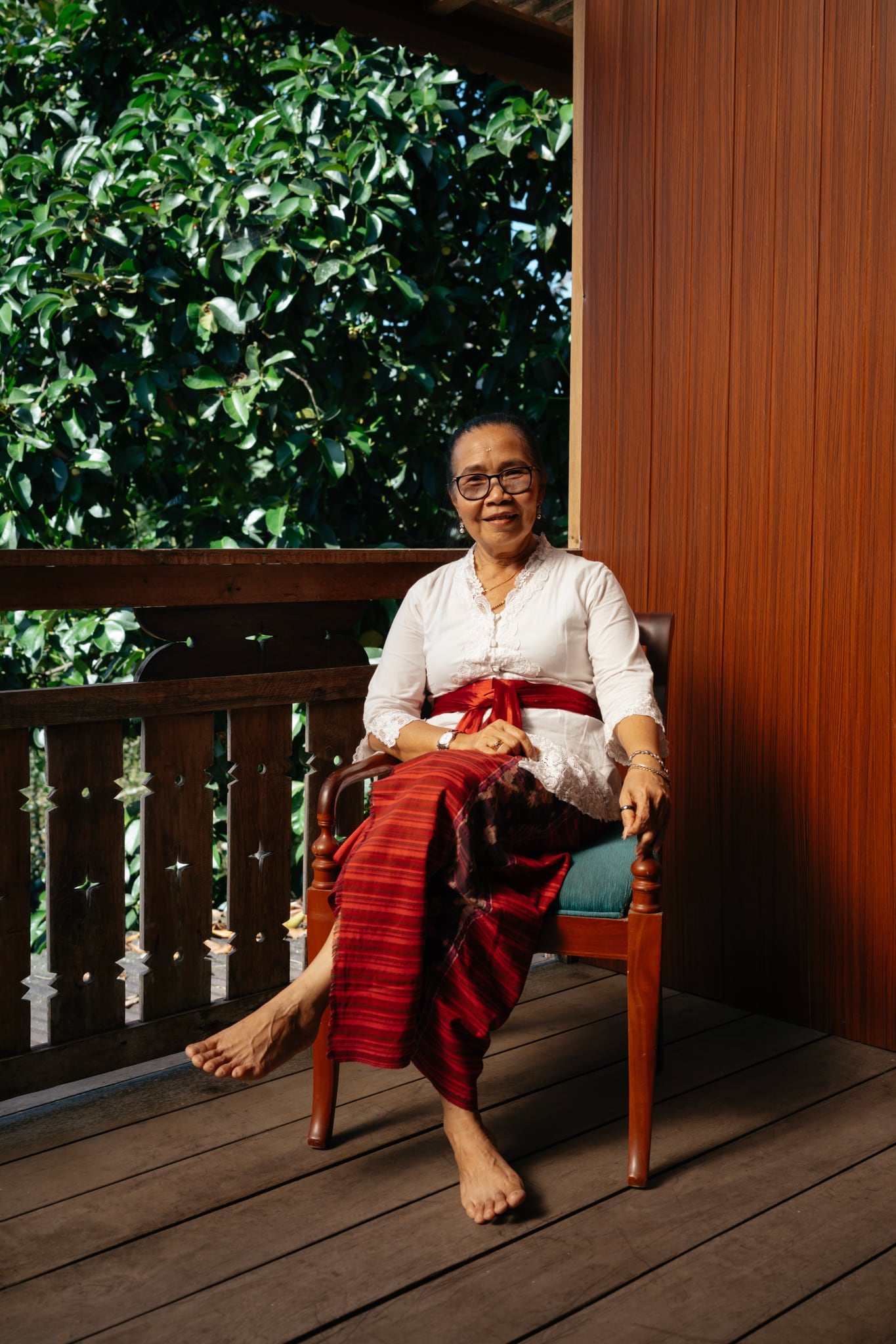 A person in a white top and red patterned cloth sits in sunlight on a wooden veranda in Bali.
