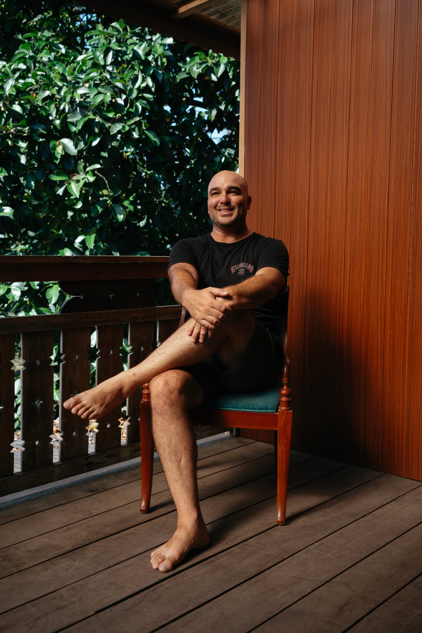 A person in dark clothing sits with crossed legs on a wooden veranda backed by green trees in Bali.