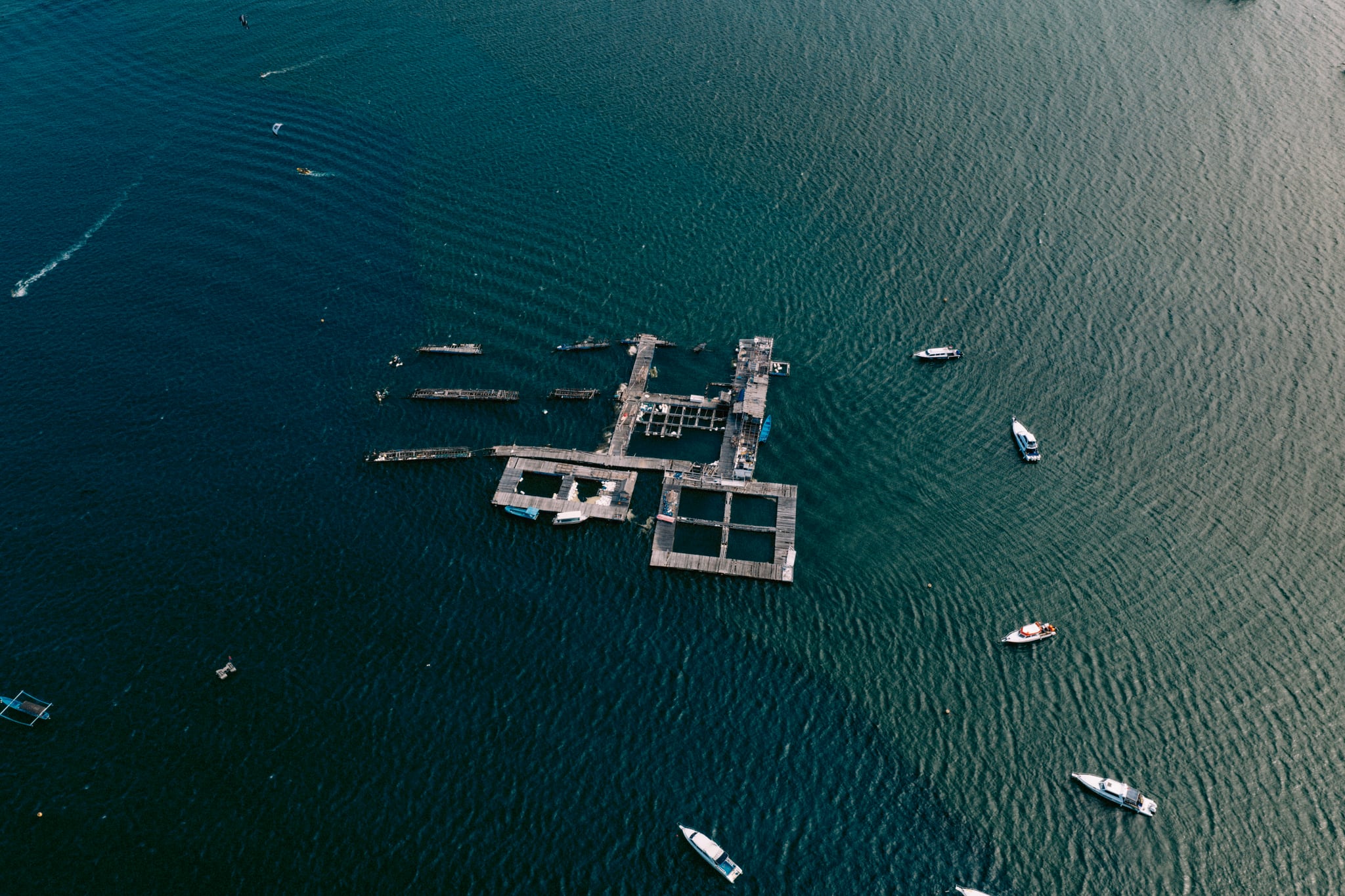 An aerial view of rectangular floating platforms and scattered boats on dark blue coastal water in Bali.