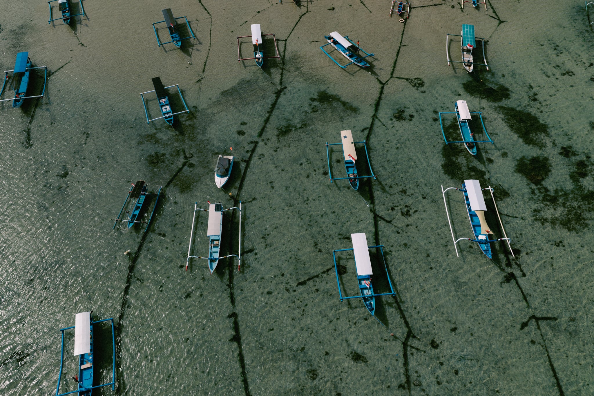 An overhead view of outrigger boats casting long shadows in shallow coastal water in Bali.
