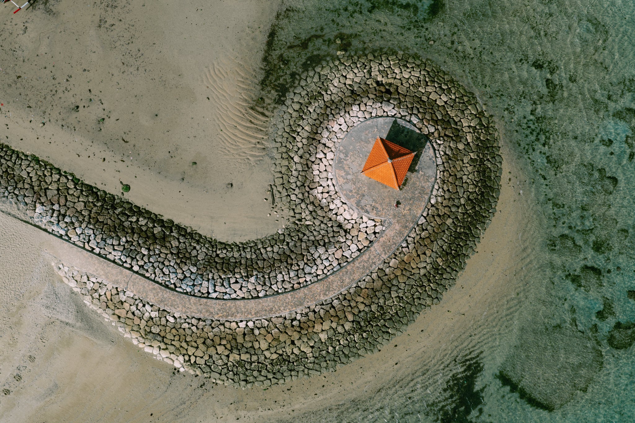 A curved stone breakwater surrounds a small pavilion with an orange roof, seen from directly above in Bali.