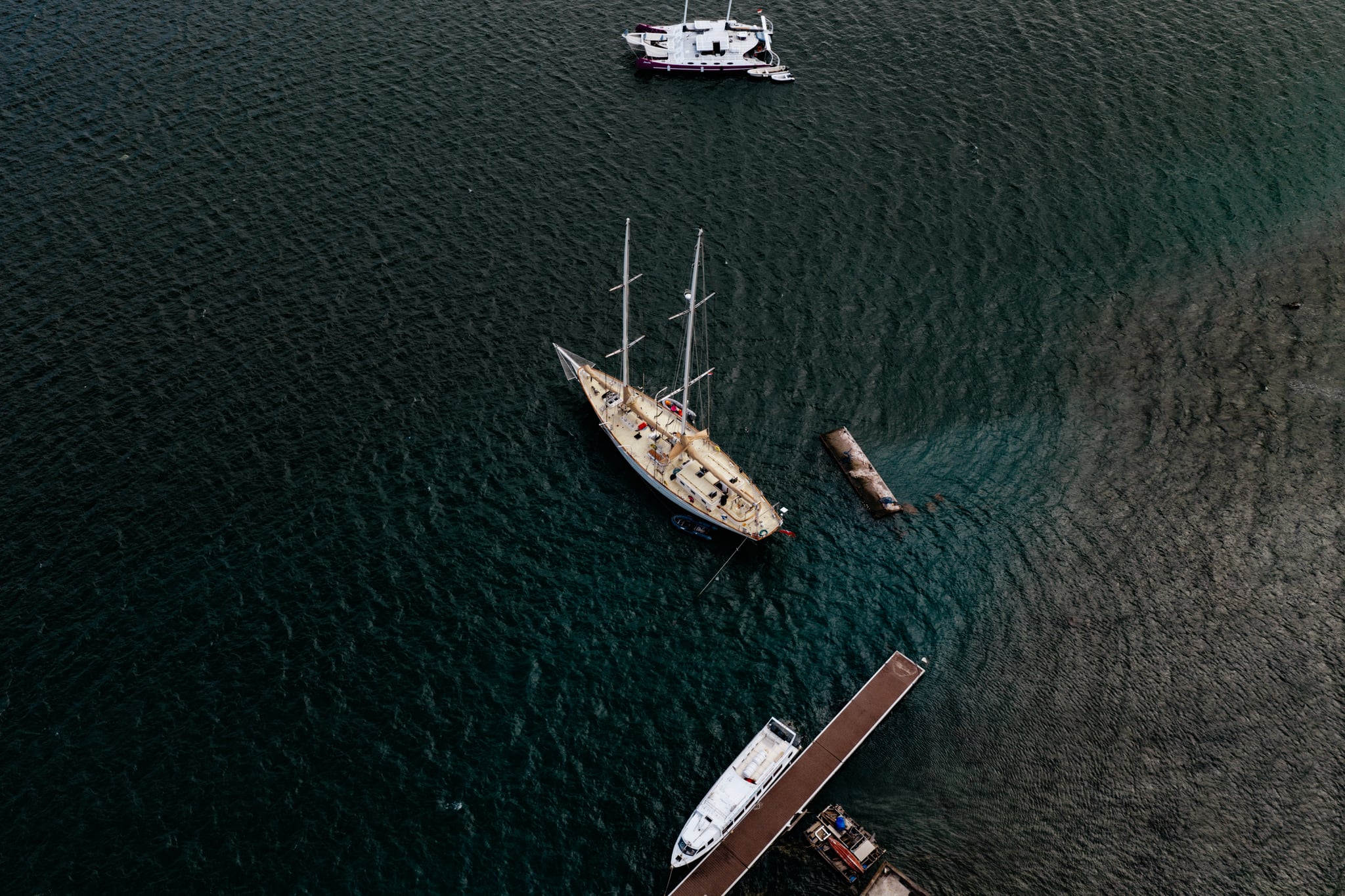 An aerial view of a wooden sailing boat, small craft, and a narrow jetty on dark coastal water in Bali.