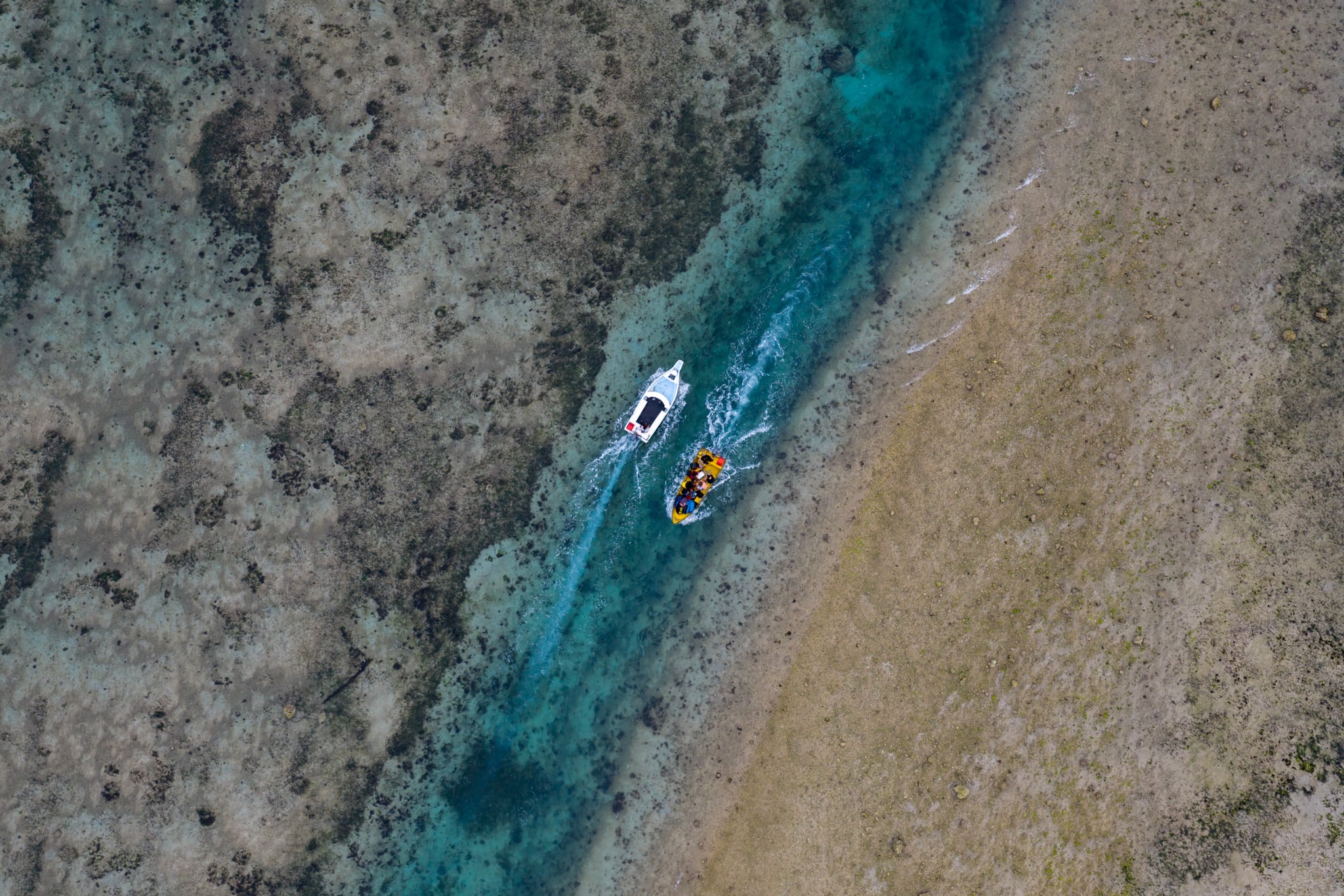 An aerial view of two small boats moving through a narrow turquoise channel between shallow reefs in Bali.