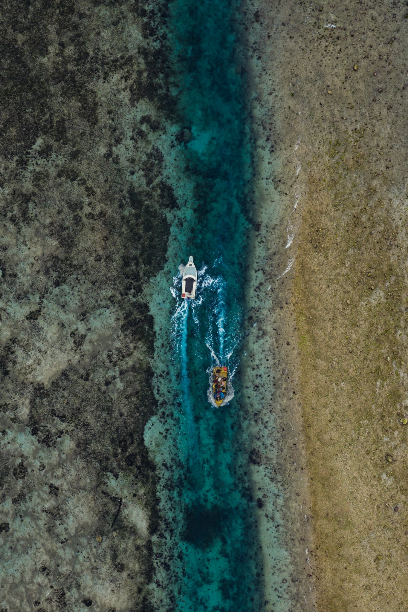 A narrow blue channel cuts through textured shallows, with small boats seen directly overhead in Bali.