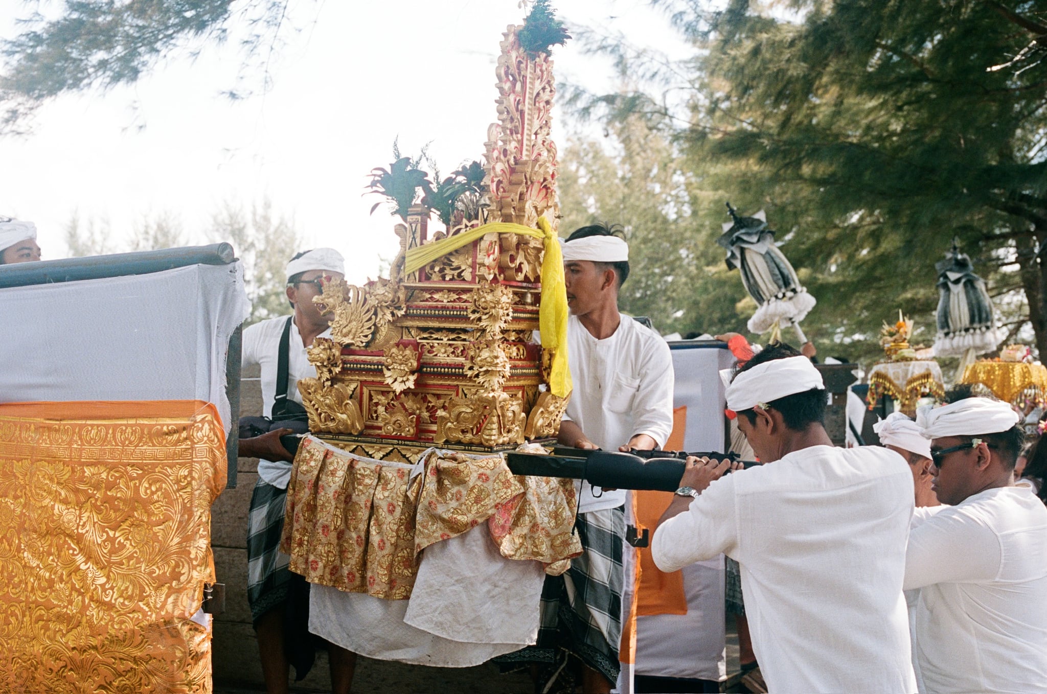 People dressed in white carry an ornate gold-coloured structure through a sunlit outdoor gathering.