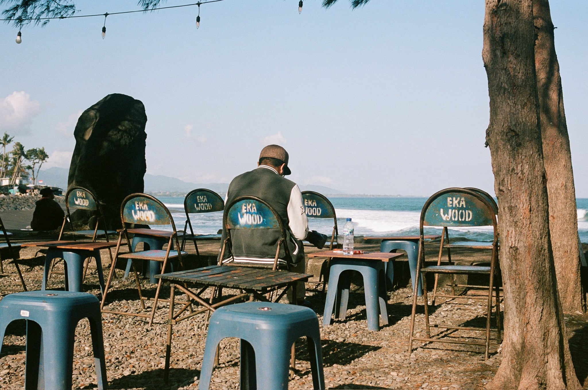 Plastic chairs and stools face the sea beneath trees, with a seated person looking toward the water.