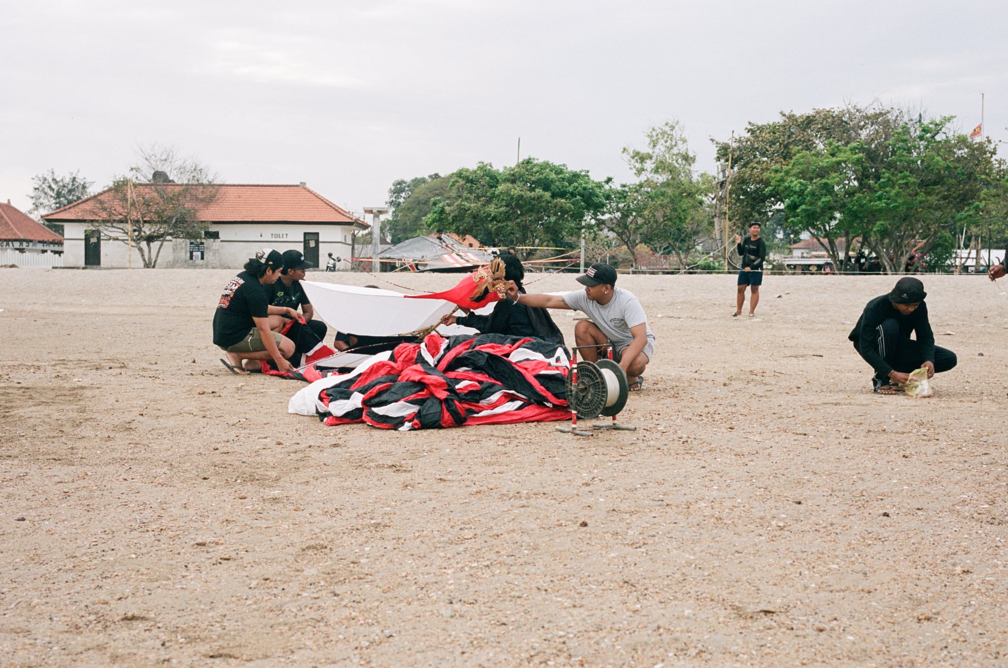 A group sits on open sandy ground around a large red, white, and black kite.