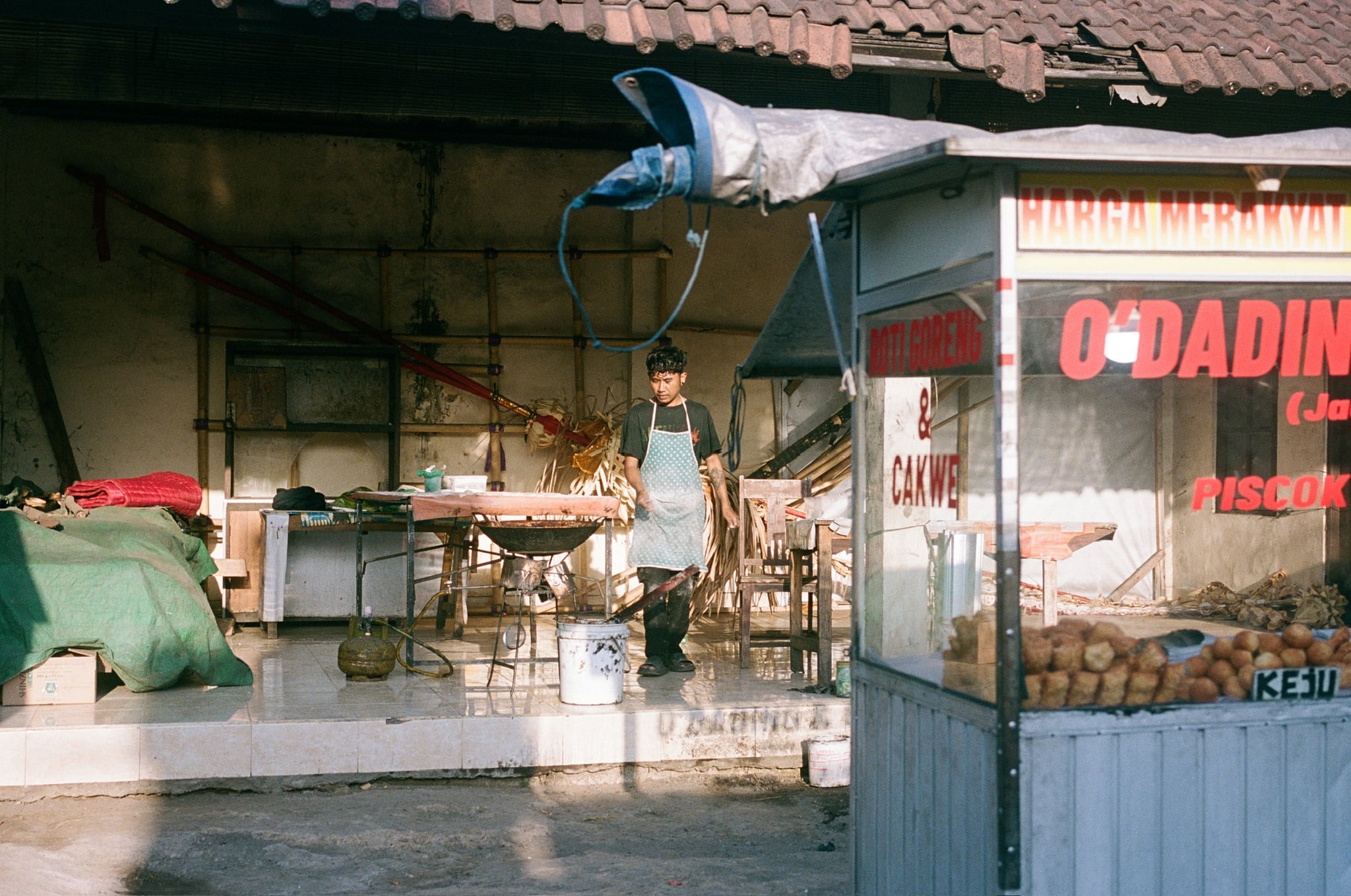 A small food stall and glass display stand beneath a tiled roof at the edge of a street.
