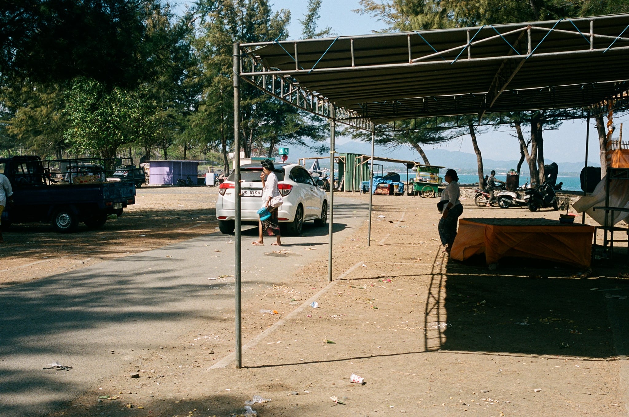 A lightweight roof shelters tables beside a broad sandy space near the sea.