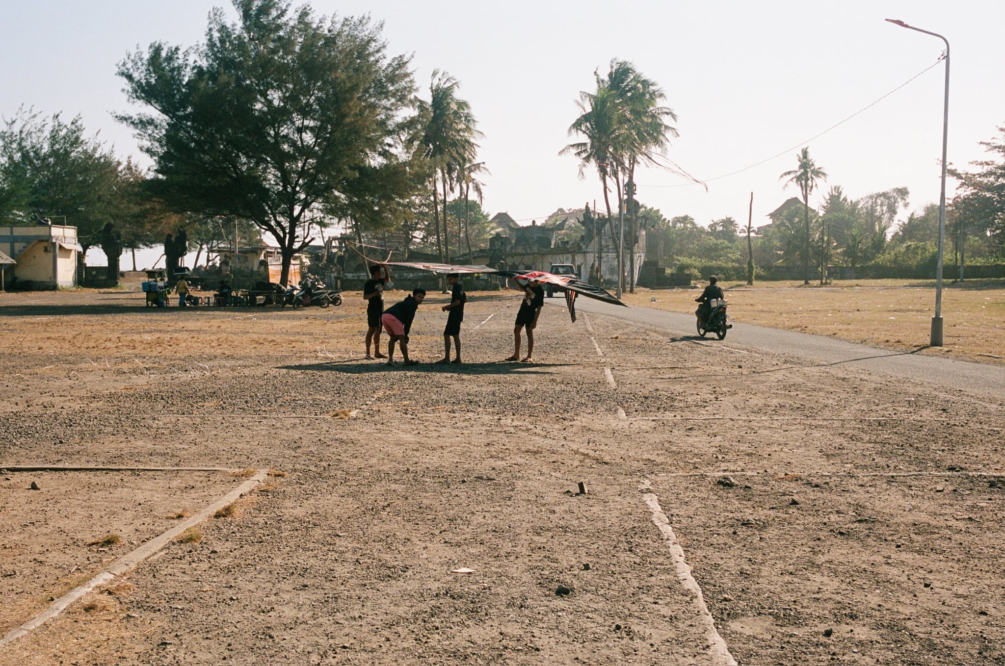 A small group stands on open ground near a road lined with palms and low buildings.