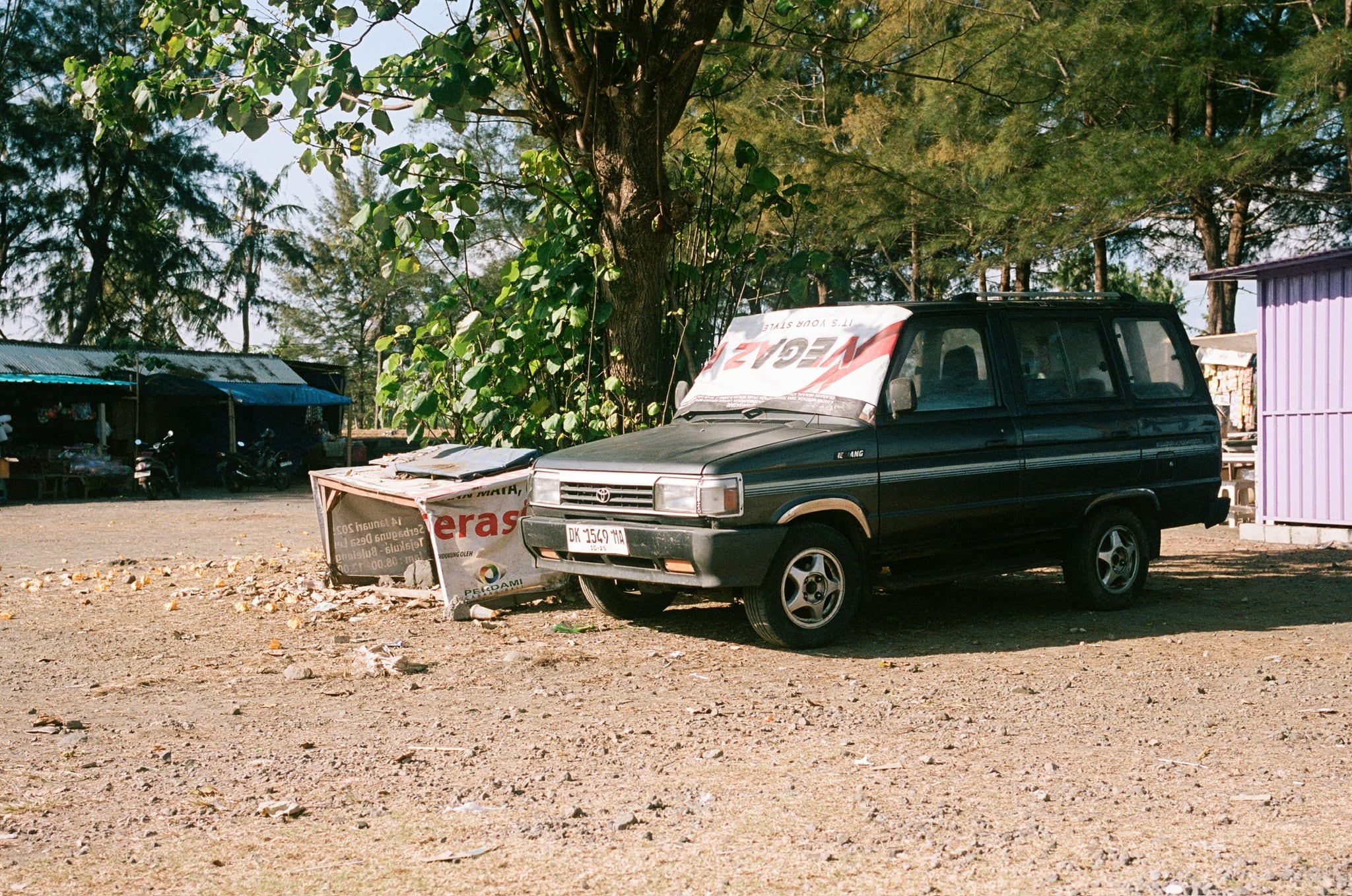 A dark green vehicle is parked beneath trees beside a roadside sign.