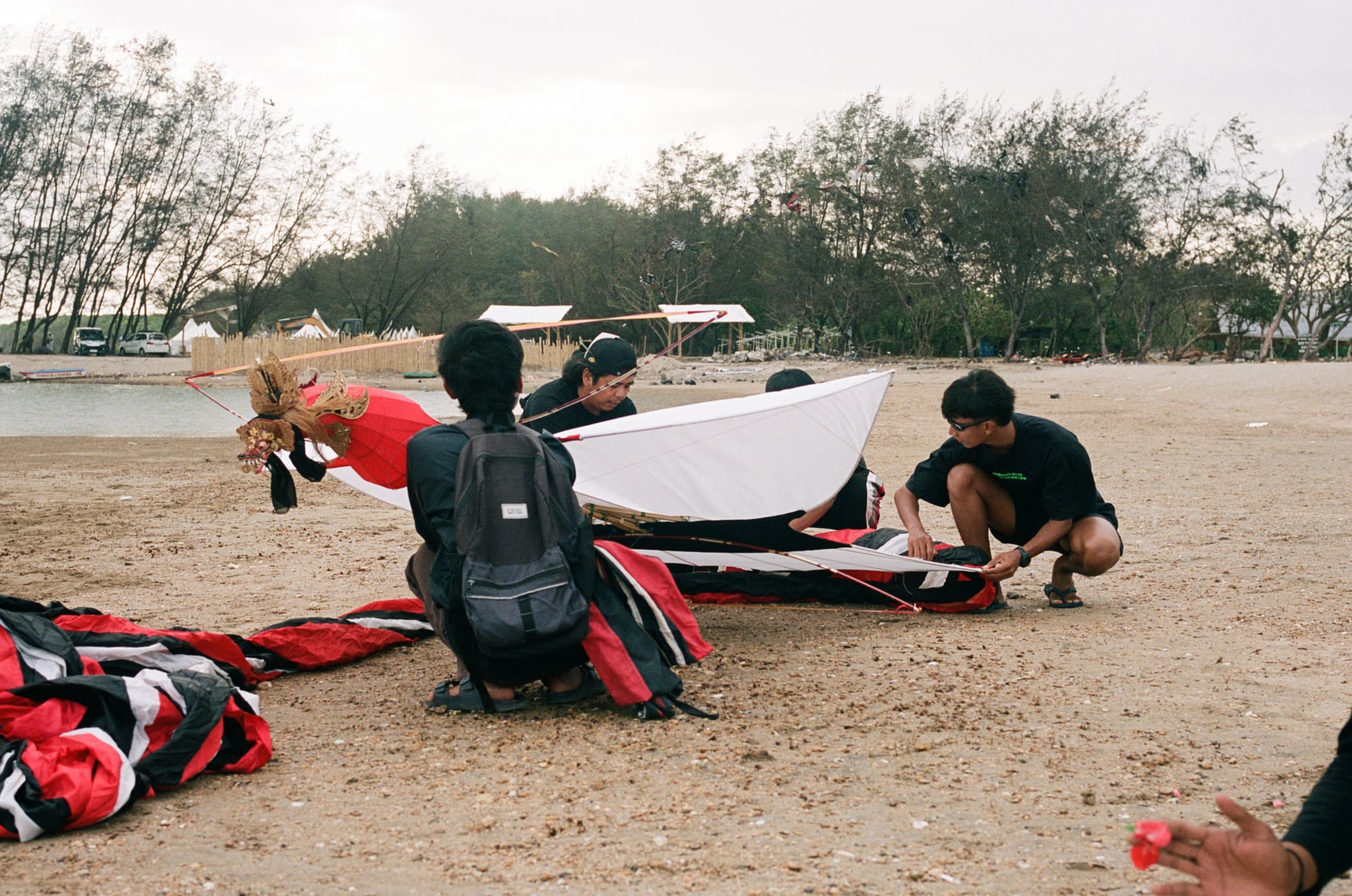 People kneel around a large red-and-white kite frame on sandy ground.