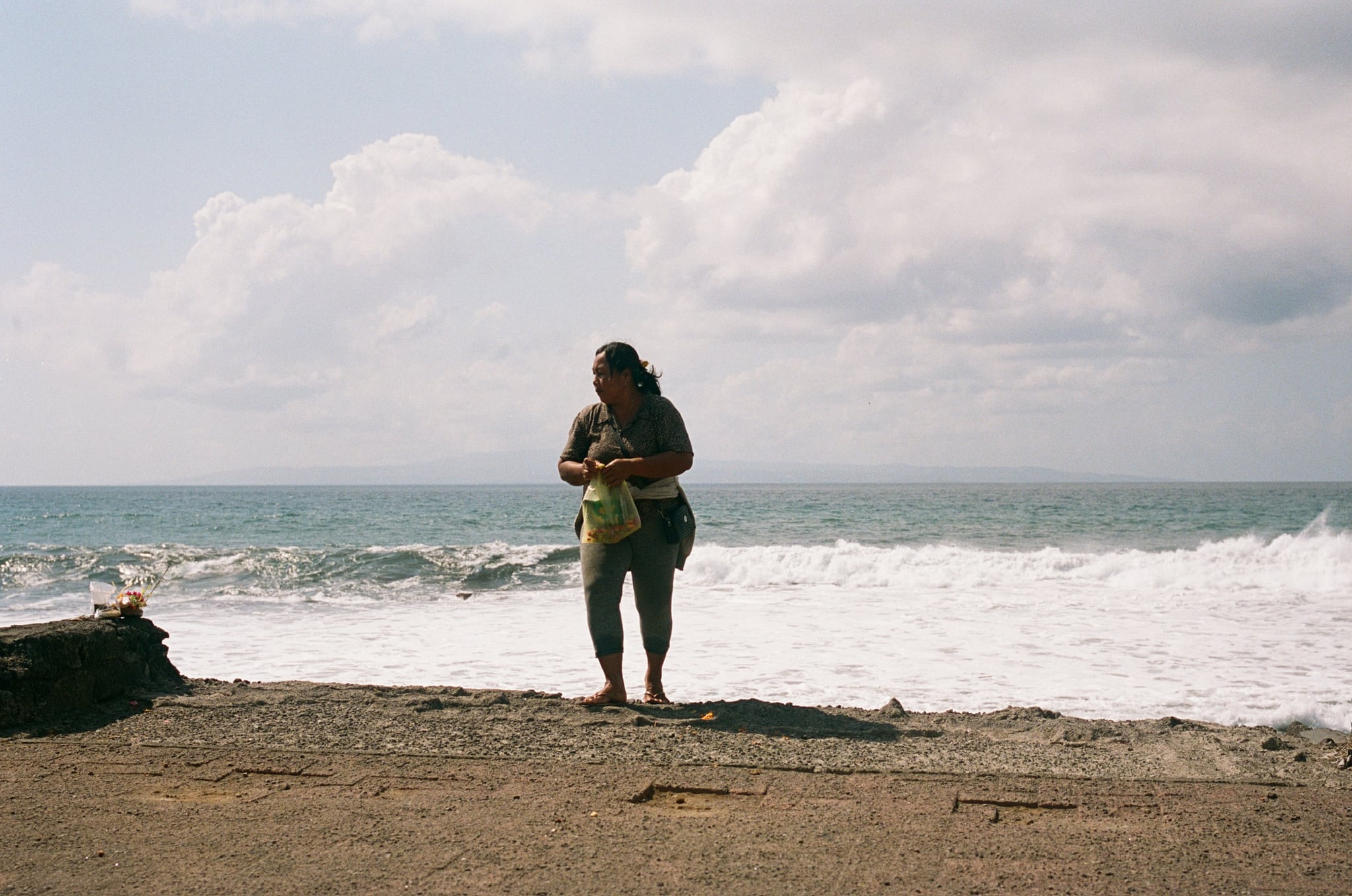 A person stands beside breaking waves beneath a bright, cloud-filled sky.
