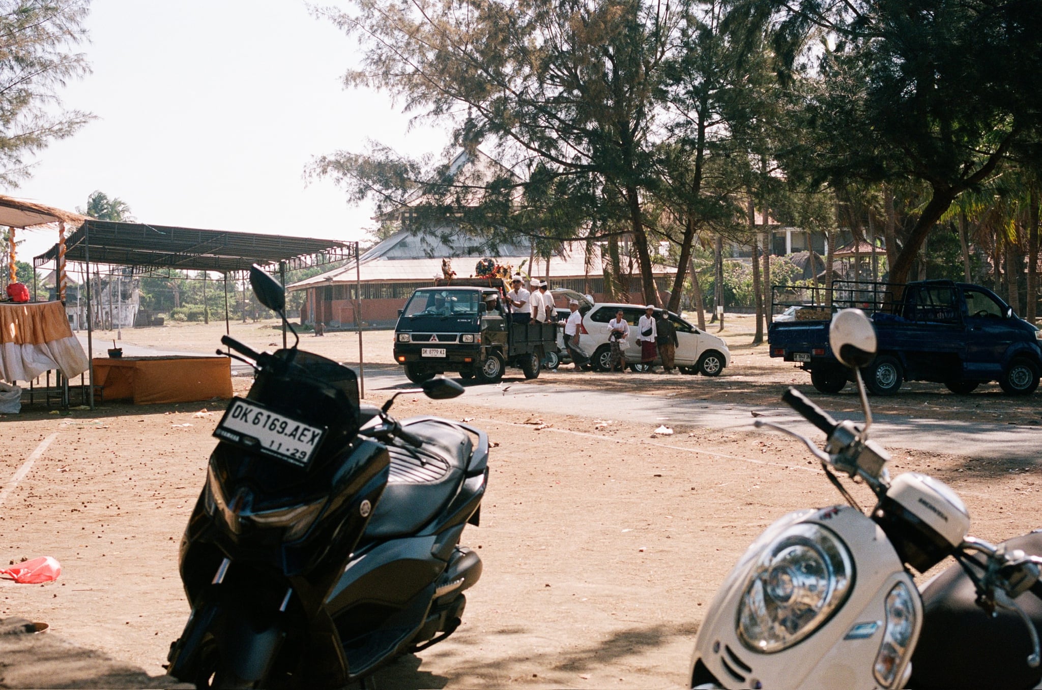 Parked scooters fill the foreground of a sandy clearing with vehicles and trees behind.