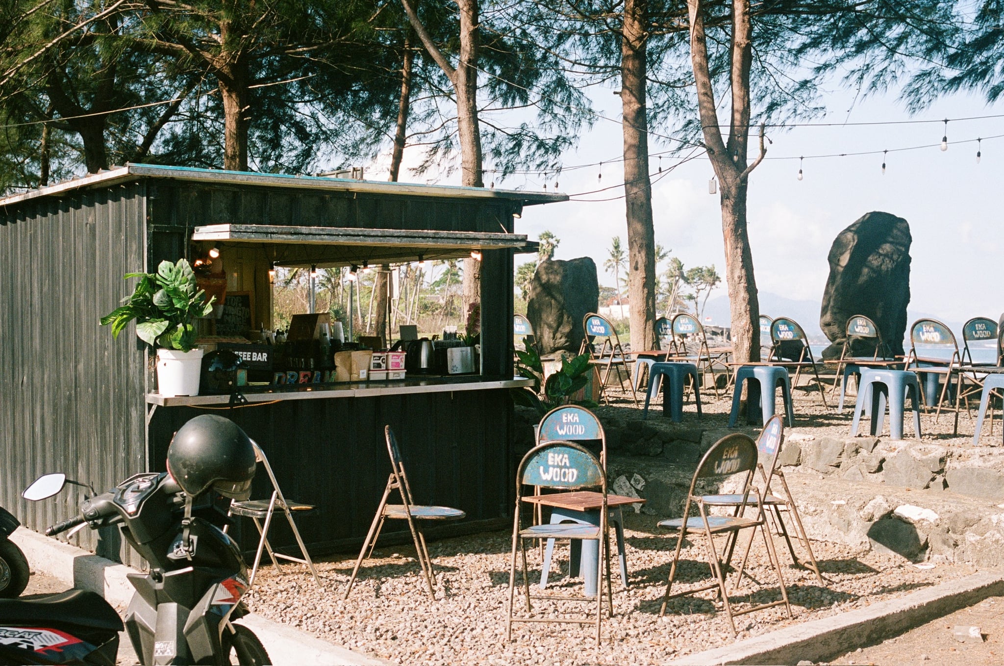 A small green kiosk stands among plastic chairs and coastal trees beside the sea.