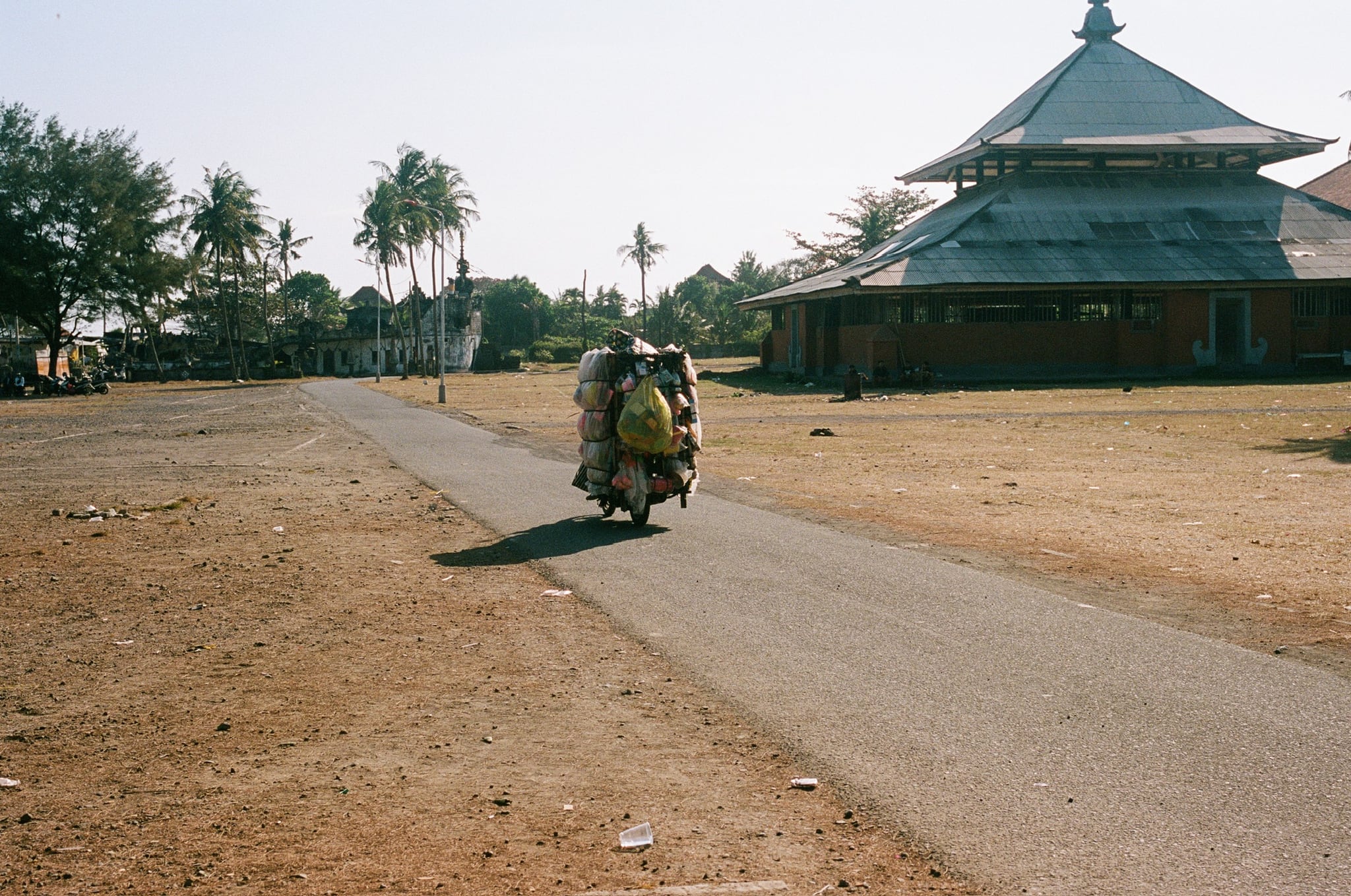 A motorcycle travels along a narrow road beside a large pavilion with a tiered roof.