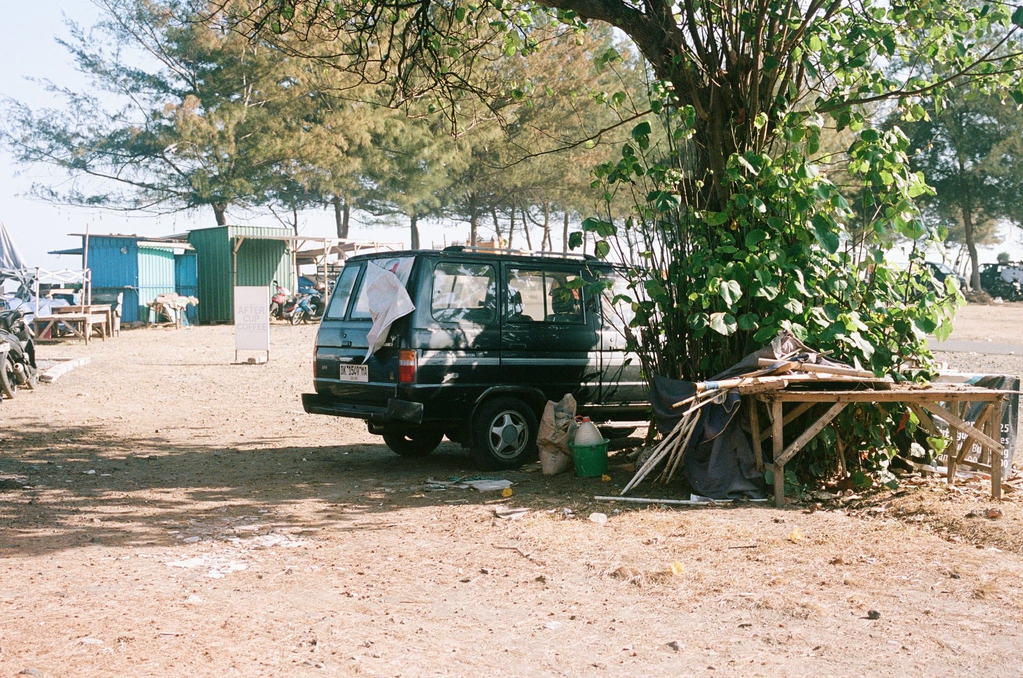 A dark vehicle is parked in dappled shade beside a low wooden platform and trees.